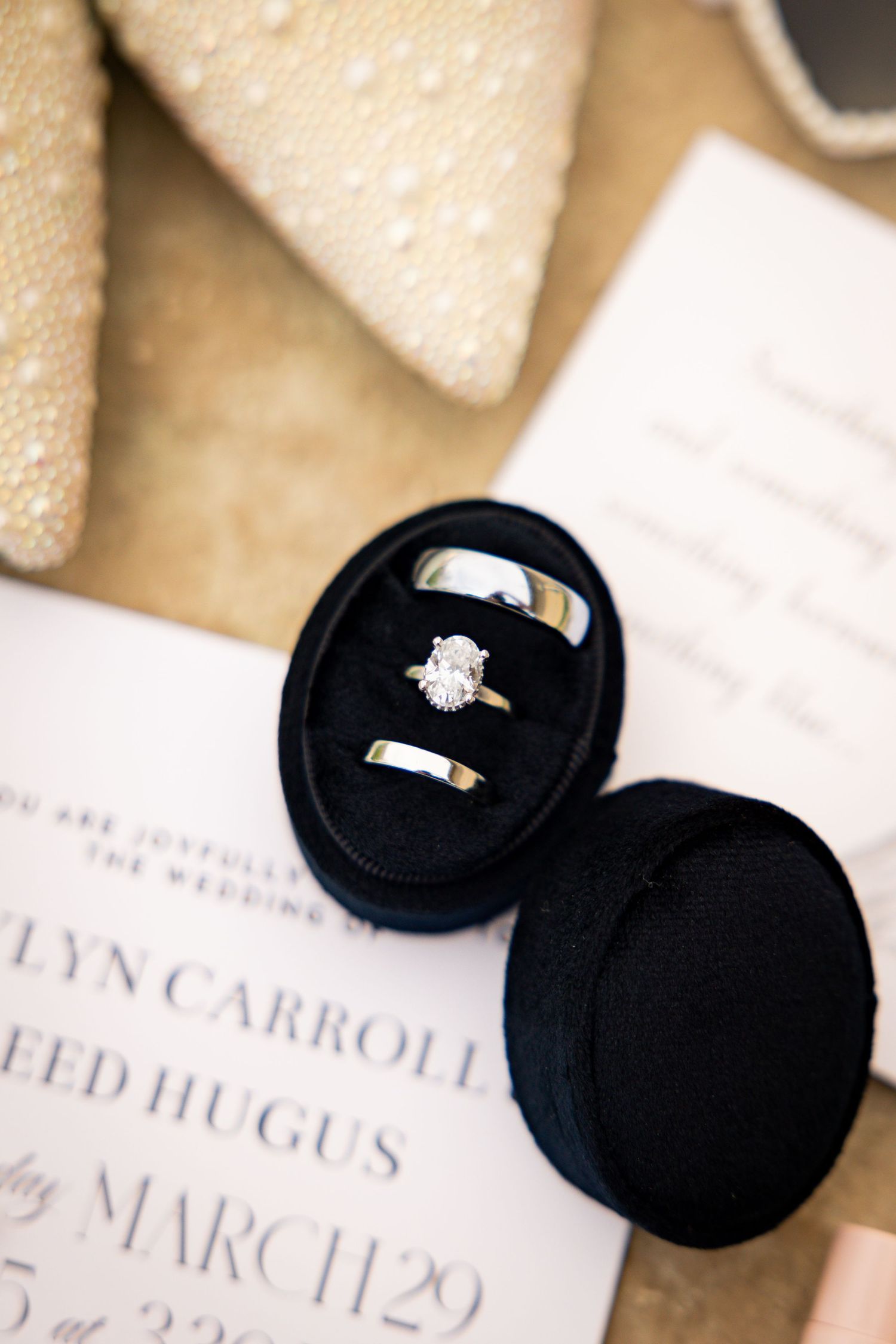 Wedding rings and engagement ring rest in black velvet ring box beside elegant ivory invitation card.