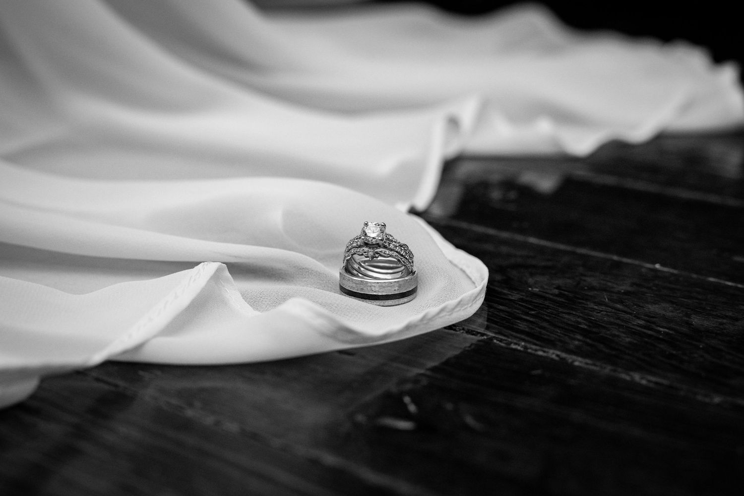 Black and white close-up of wedding rings on a wooden surface with soft fabric in the background at Harvest Adventures wedding venue.