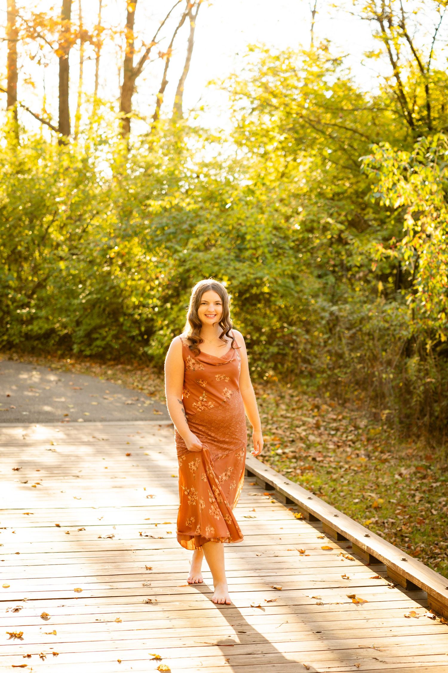 A woman in a rust-colored dress poses on a wooden boardwalk surrounded by autumn foliage.