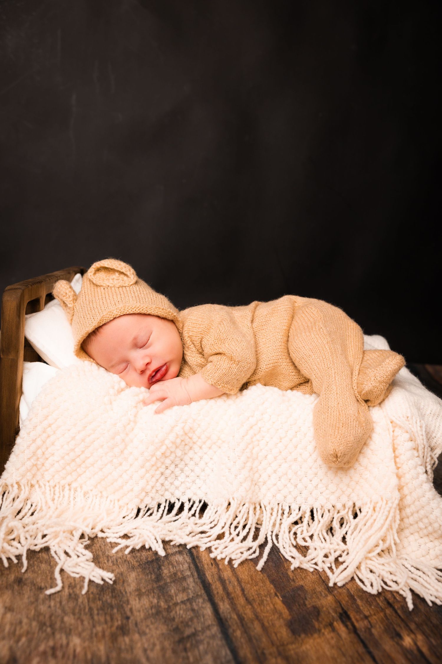 Sleeping newborn wearing cozy beige knit outfit rests peacefully on white fringed blanket atop wooden surface.