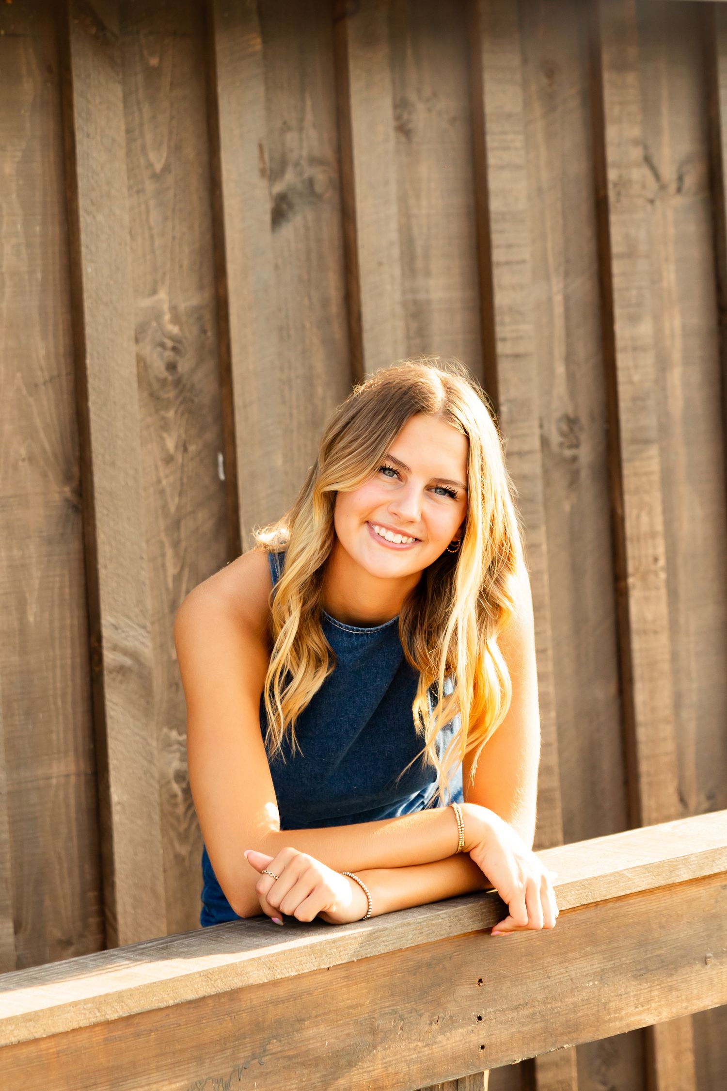Young blonde woman in denim top smiling while leaning on wooden fence railing outdoors.