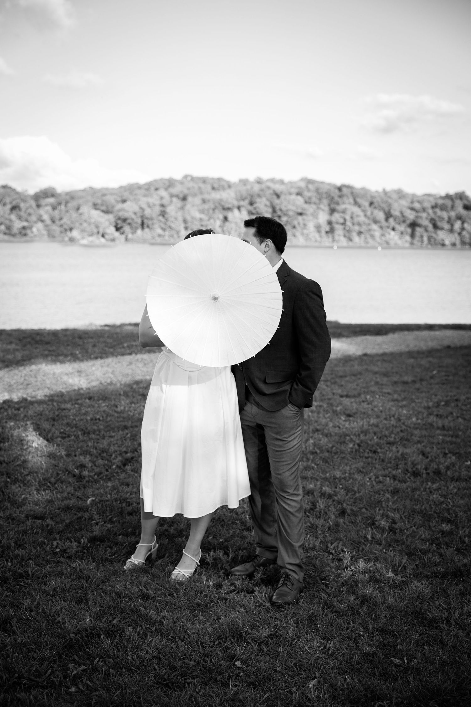 Black and white artistic wedding photograph showing couple embracing in a scenic meadow with hills in background.
