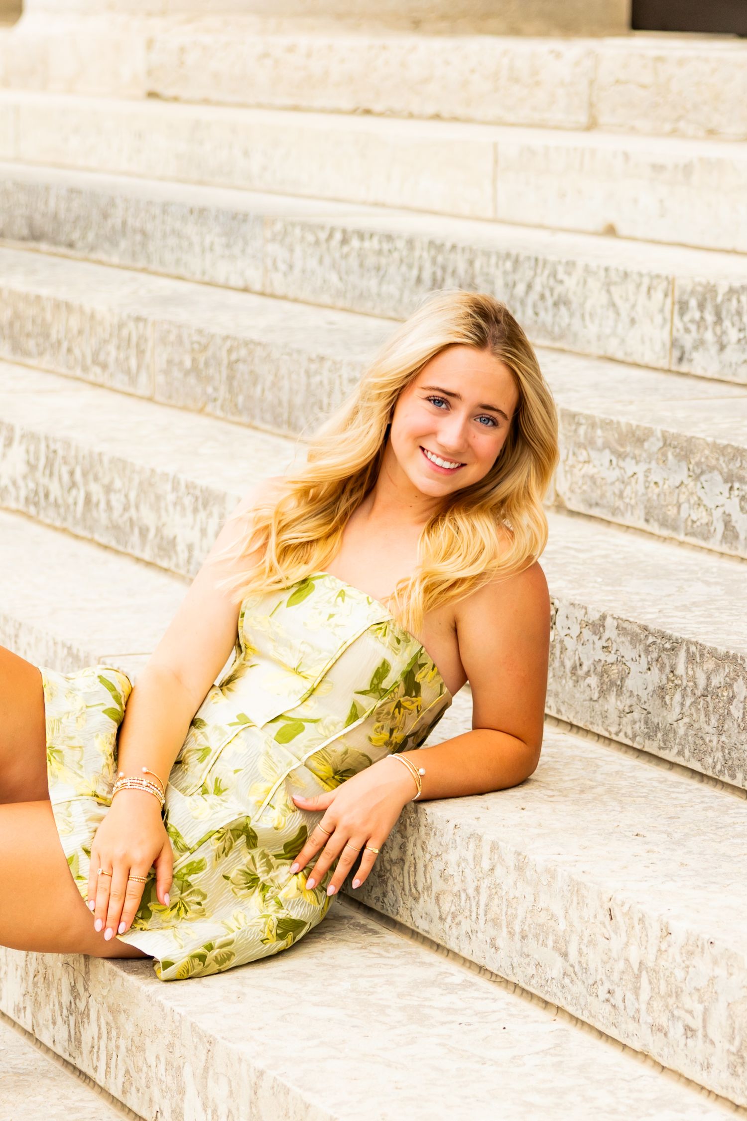 Young blonde woman in a floral sundress sitting on marble steps, smiling outdoors in warm sunlight.