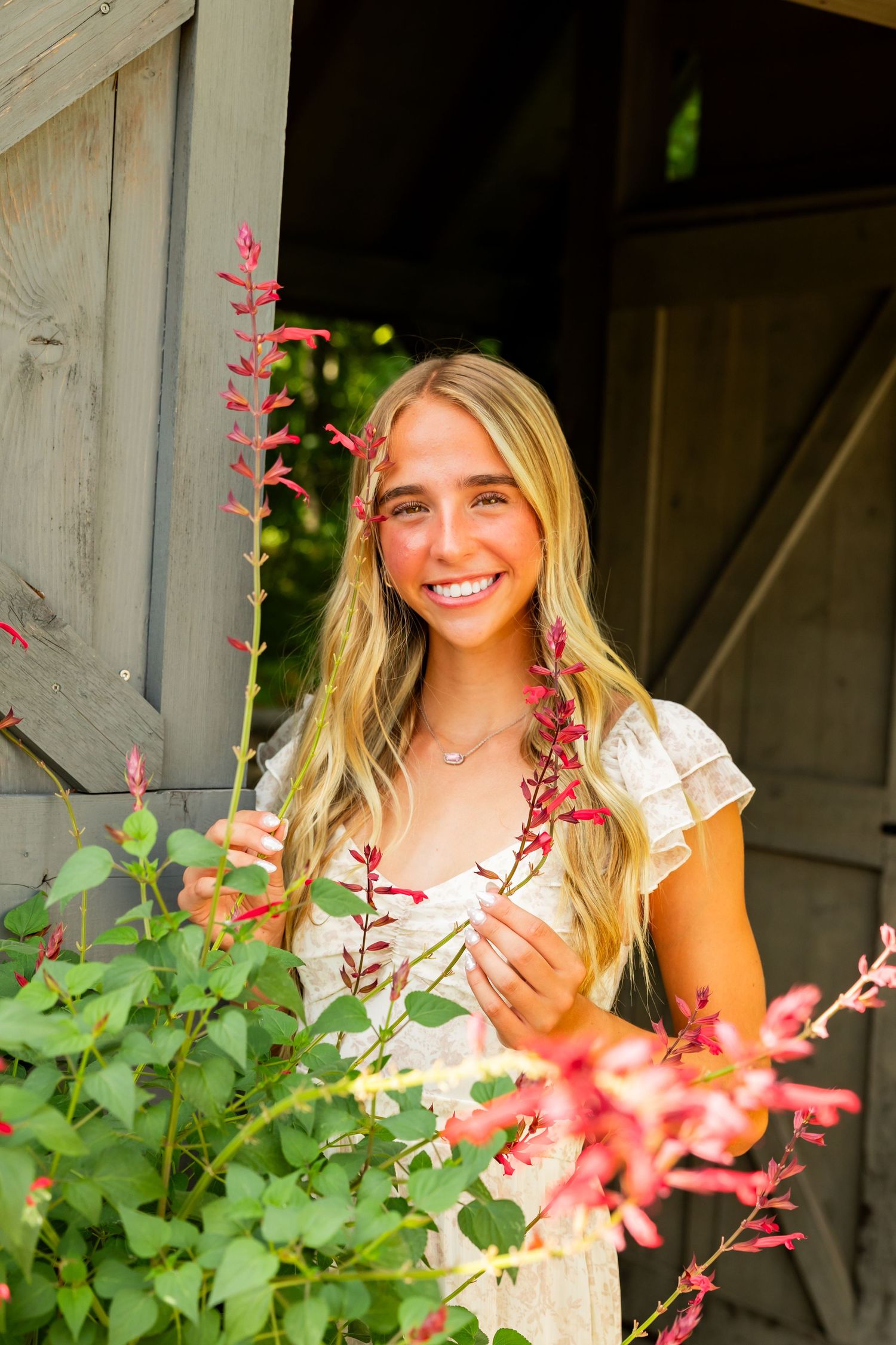 Smiling blonde teenage girl in white dress holding pink flowers near rustic wooden barn door outdoors.