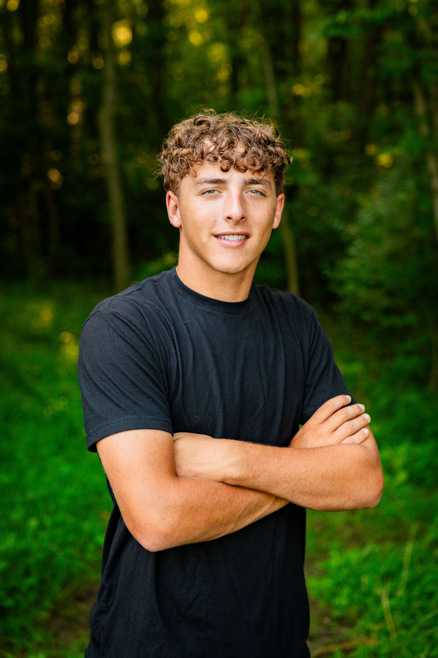 Person in black t-shirt stands with arms crossed in front of green forest background.