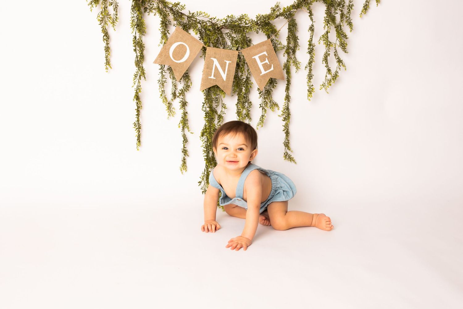 Sweet baby lies on white backdrop beneath hanging greenery garland during milestone photo session.