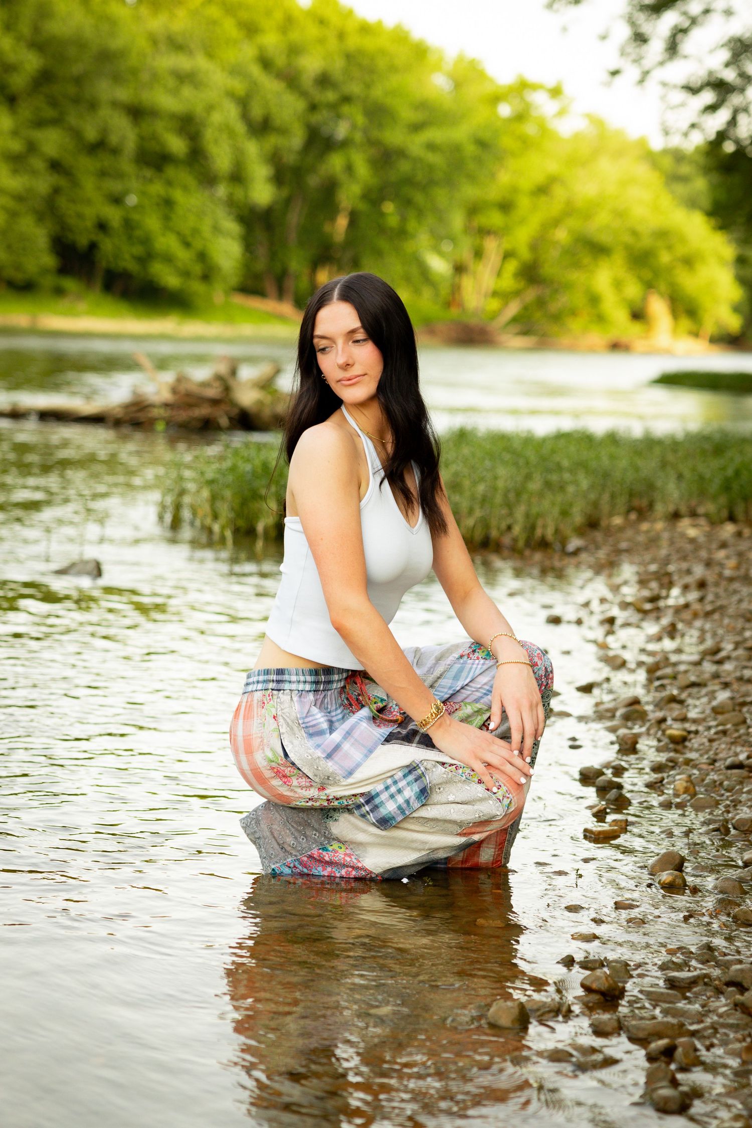 A person in a white tank top and plaid shorts poses by the water's edge on a rocky riverbank in summer.