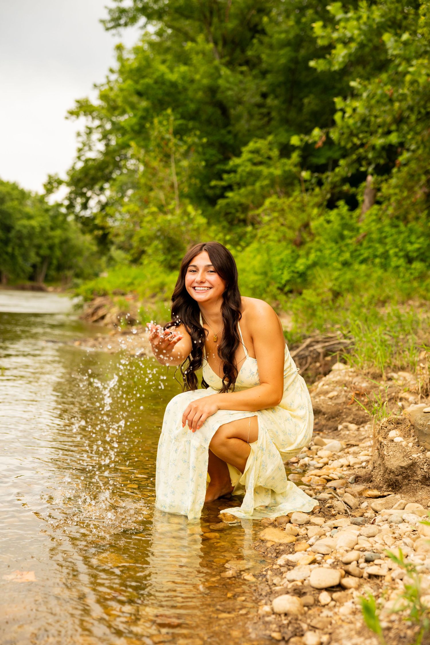 A smiling woman in a white dress crouches by a rocky stream, splashing water with her hands, surrounded by lush green trees.