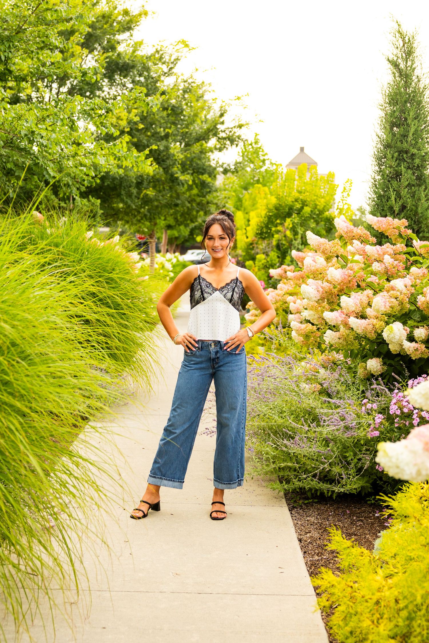 Woman in white lace top and wide-leg jeans poses on garden path surrounded by hydrangeas and ornamental grasses.