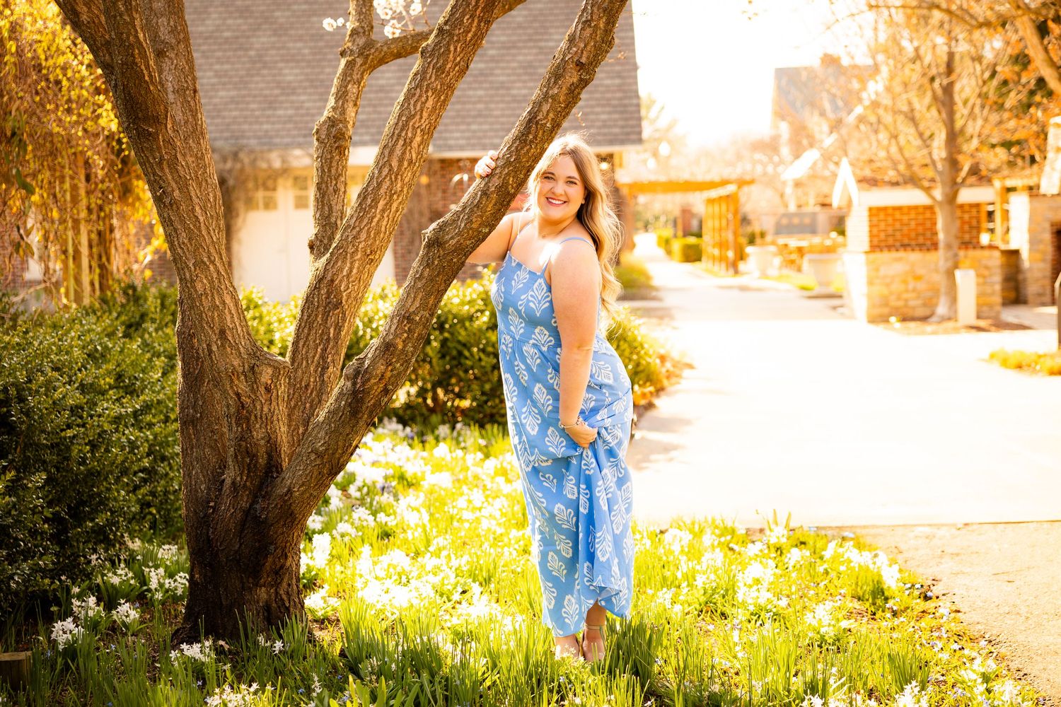 A person in a blue dress walks through a sunlit garden path with blooming daffodils and trees.
