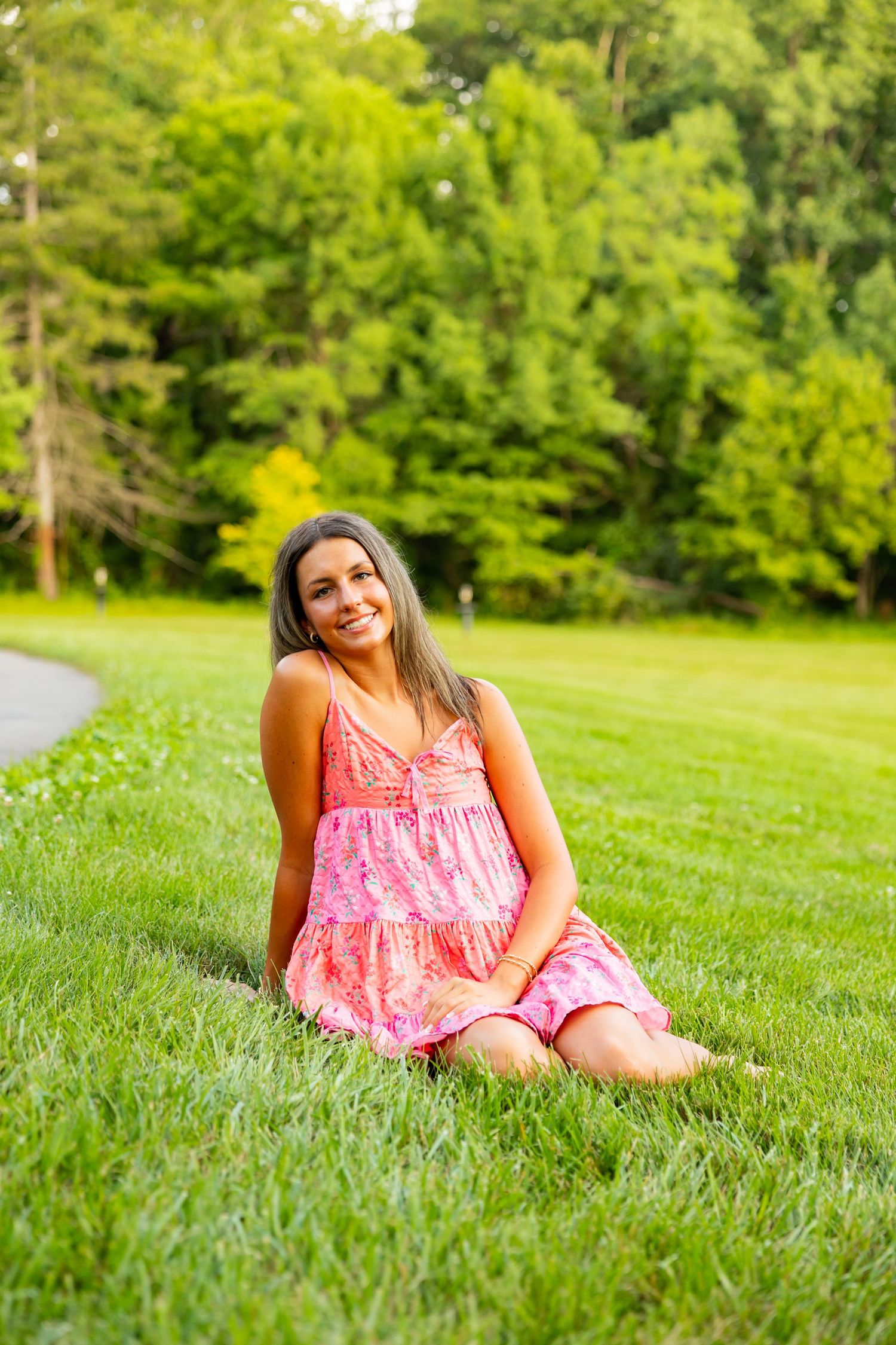 Young woman in pink sundress sitting on lush green grass, smiling outdoors with trees in background.
