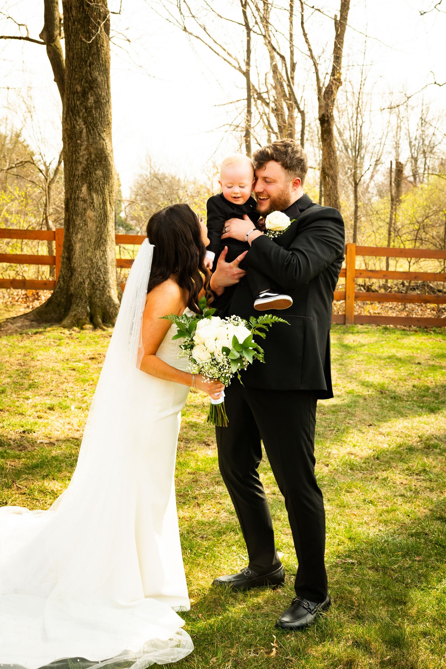 Newlyweds share an intimate outdoor moment with their baby in a rustic garden setting with white bridal bouquet.
