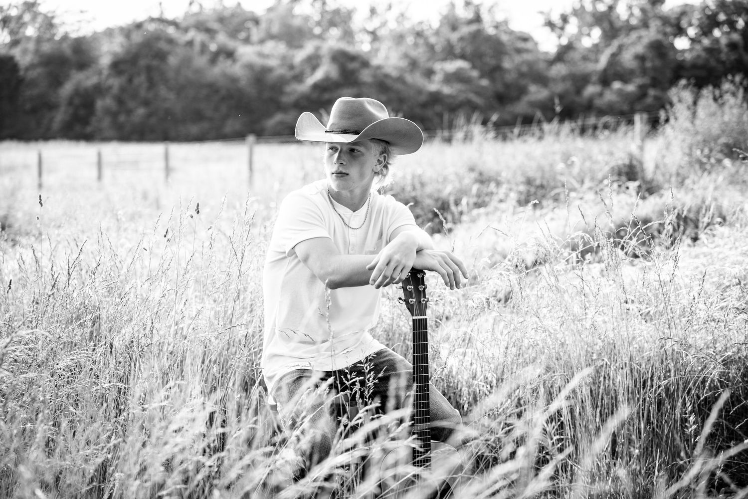 Black and white photograph of someone sitting in tall grass on a hillside wearing a hat and casual attire.