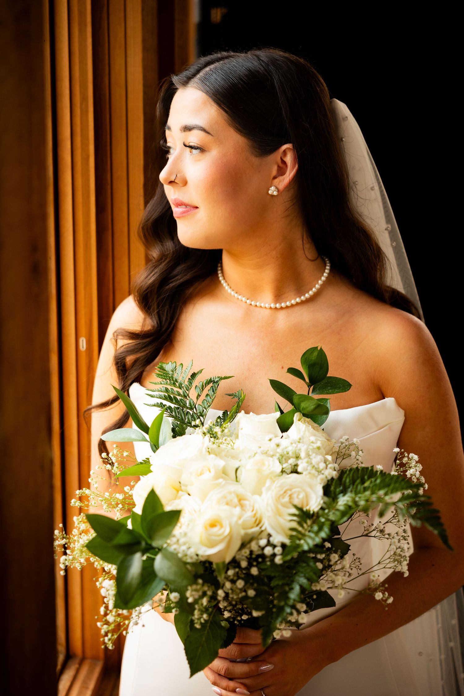 Elegant bride in strapless gown holds white rose bouquet with greenery against golden curtained backdrop.