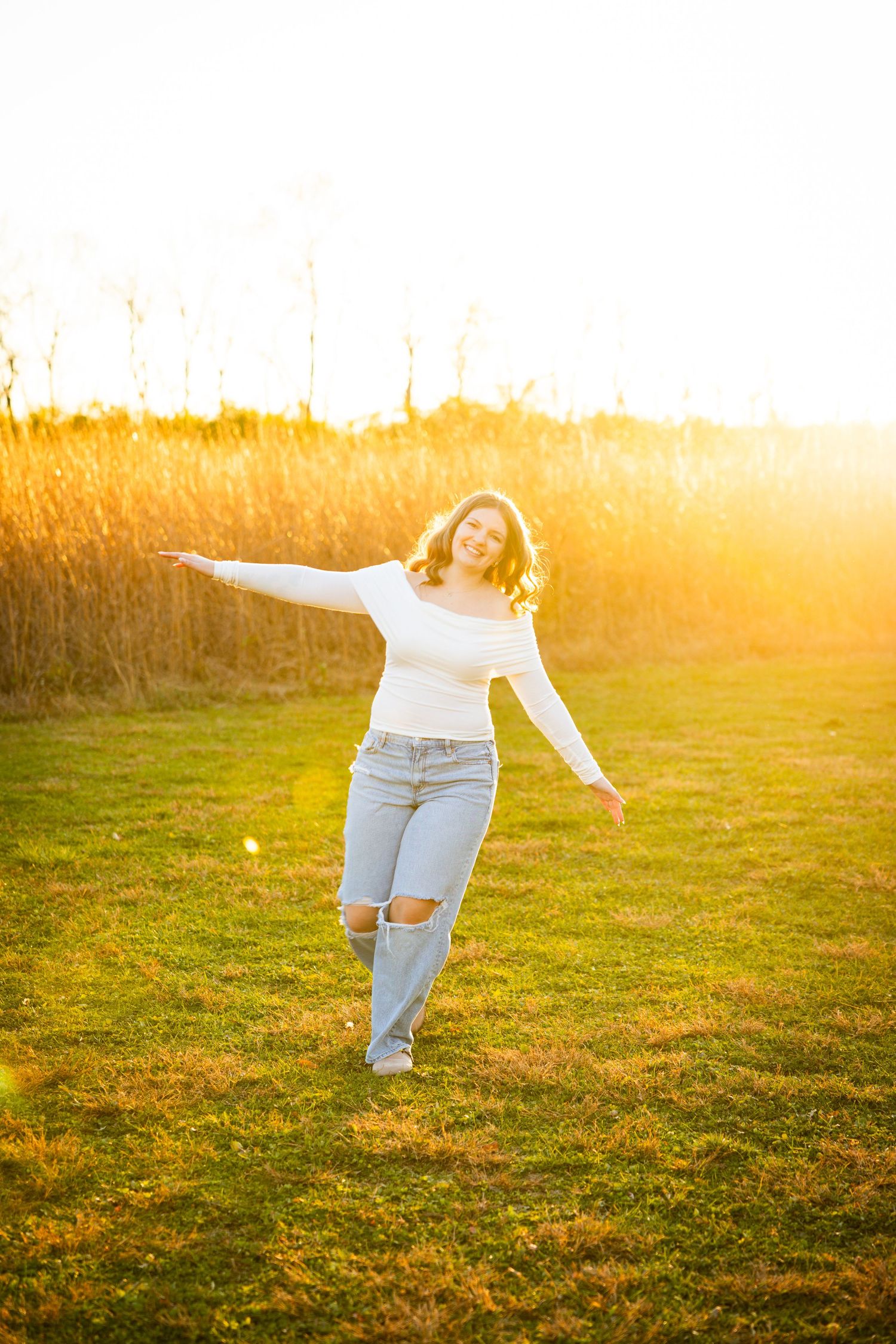 A person in ripped jeans and white top dances joyfully in a golden sunlit field during sunset.