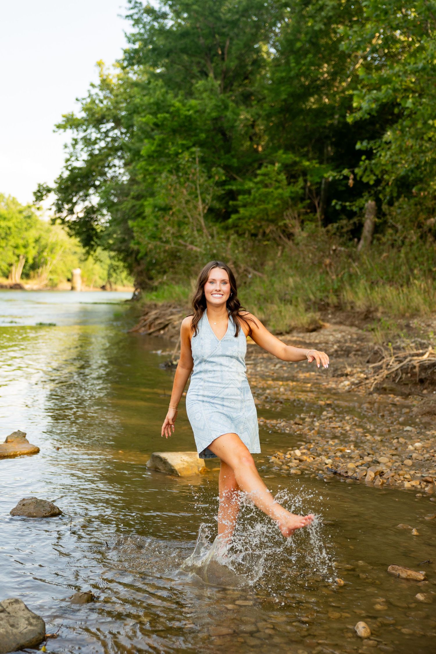 Young woman in light blue dress splashing water in a rocky river, smiling joyfully with green trees in background.