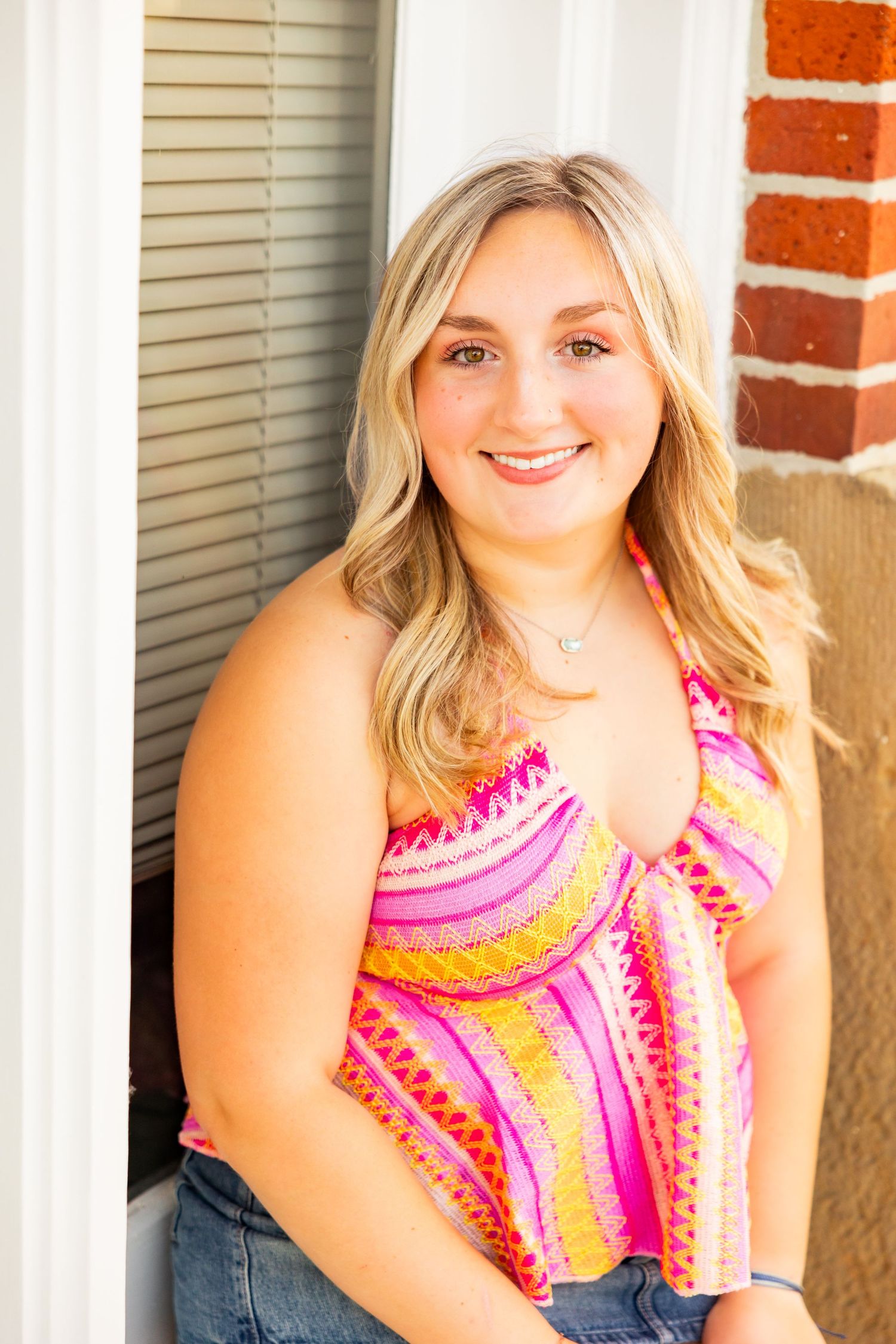 Smiling young blonde woman in colorful striped halter top and jeans leaning against white door frame.