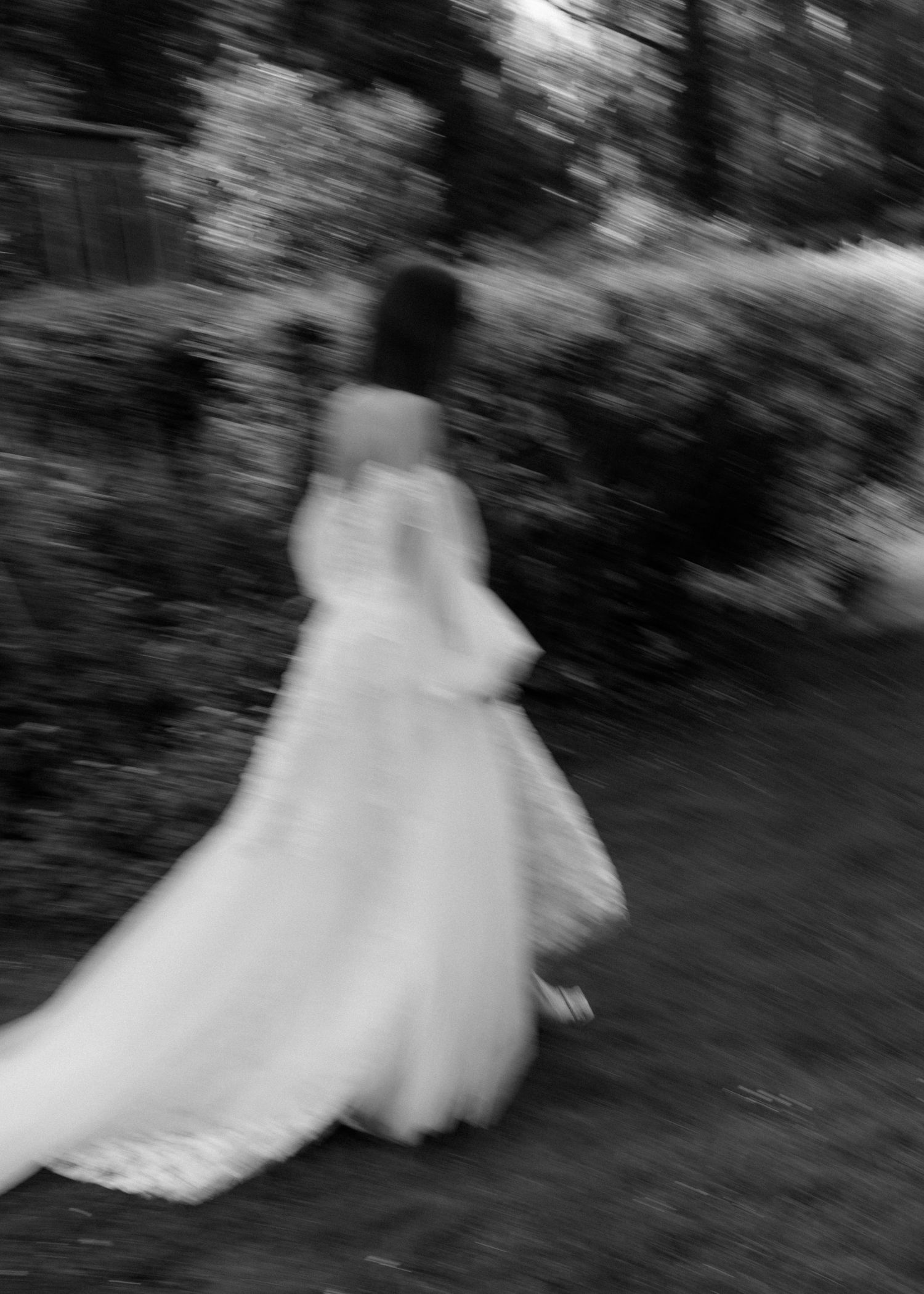 Zwart-wit motion blur foto van iemand in een vloeiende trouwjurk die door een tuinpad loopt.