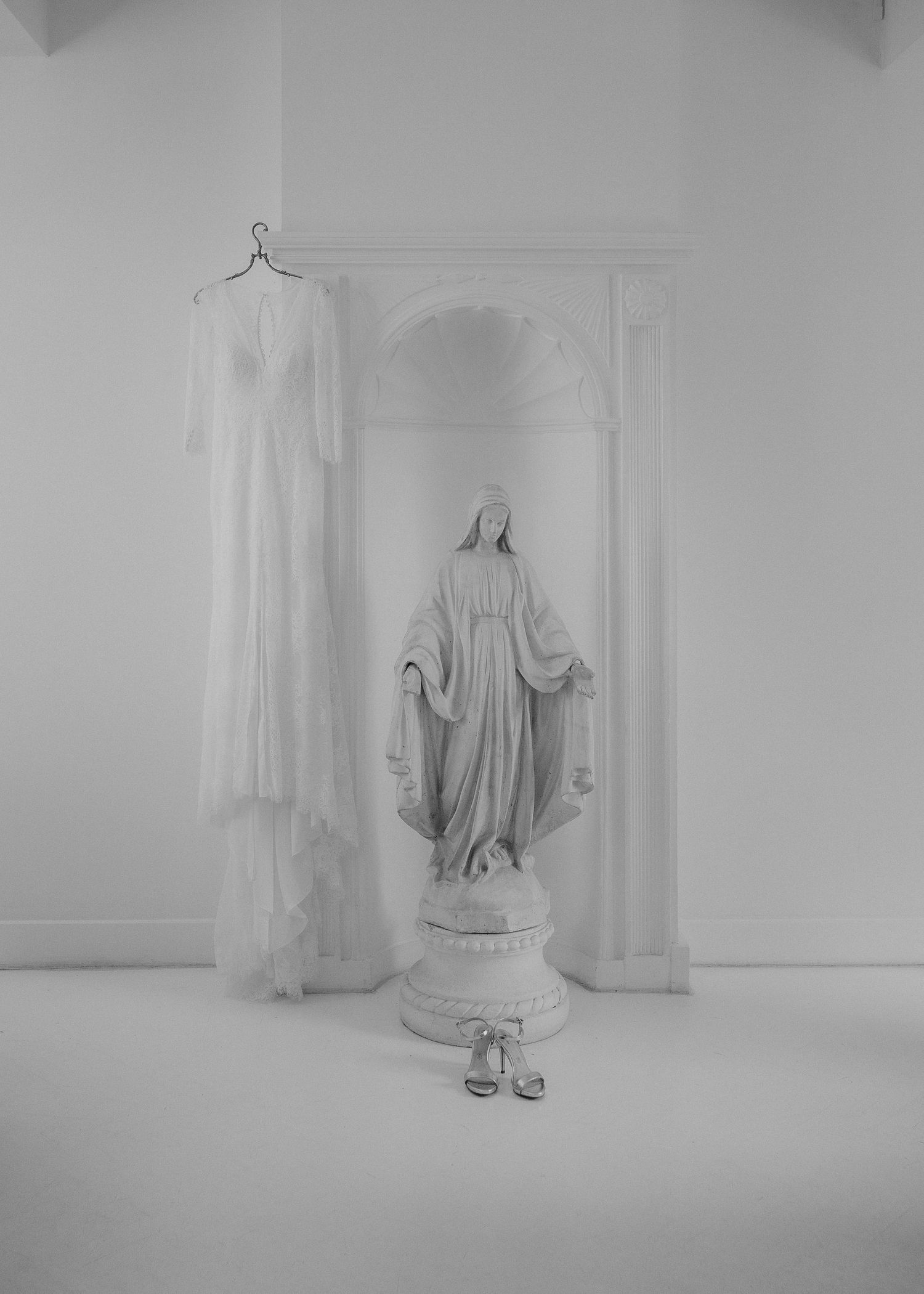 Witte trouwjurk hangt naast een Mariabeeld in een serene, lichte ruimte, in kasteel hoogenweerth met bruidsschoenen ervoor geplaatst