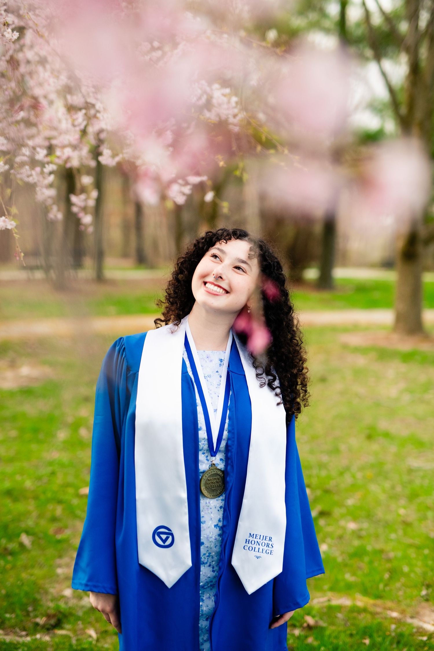 Graduate in blue cap and gown with white stole stands smiling under blooming cherry blossoms in spring.