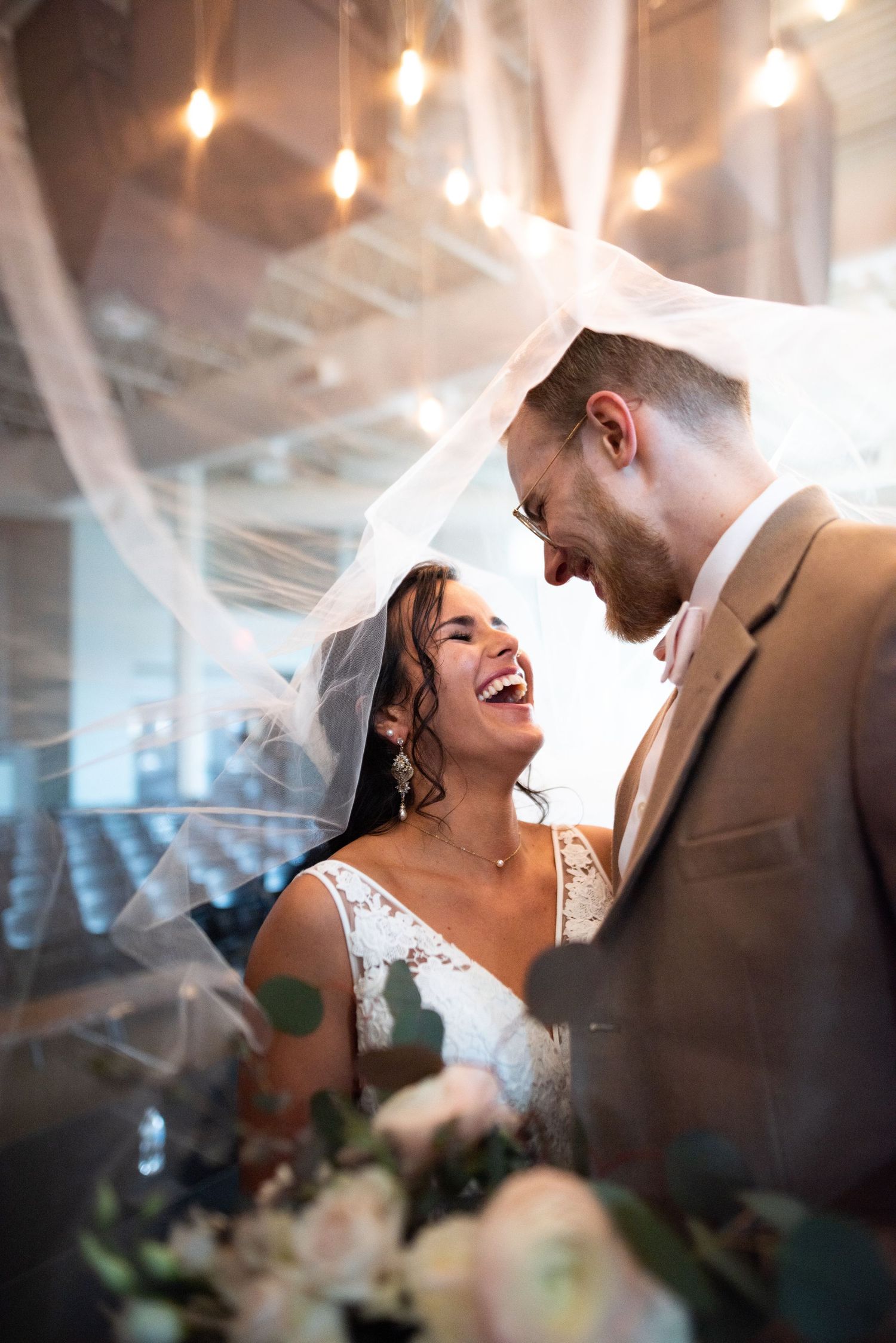 Newlywed couple shares joyful laughter under a veil with string lights glowing in an indoor venue.