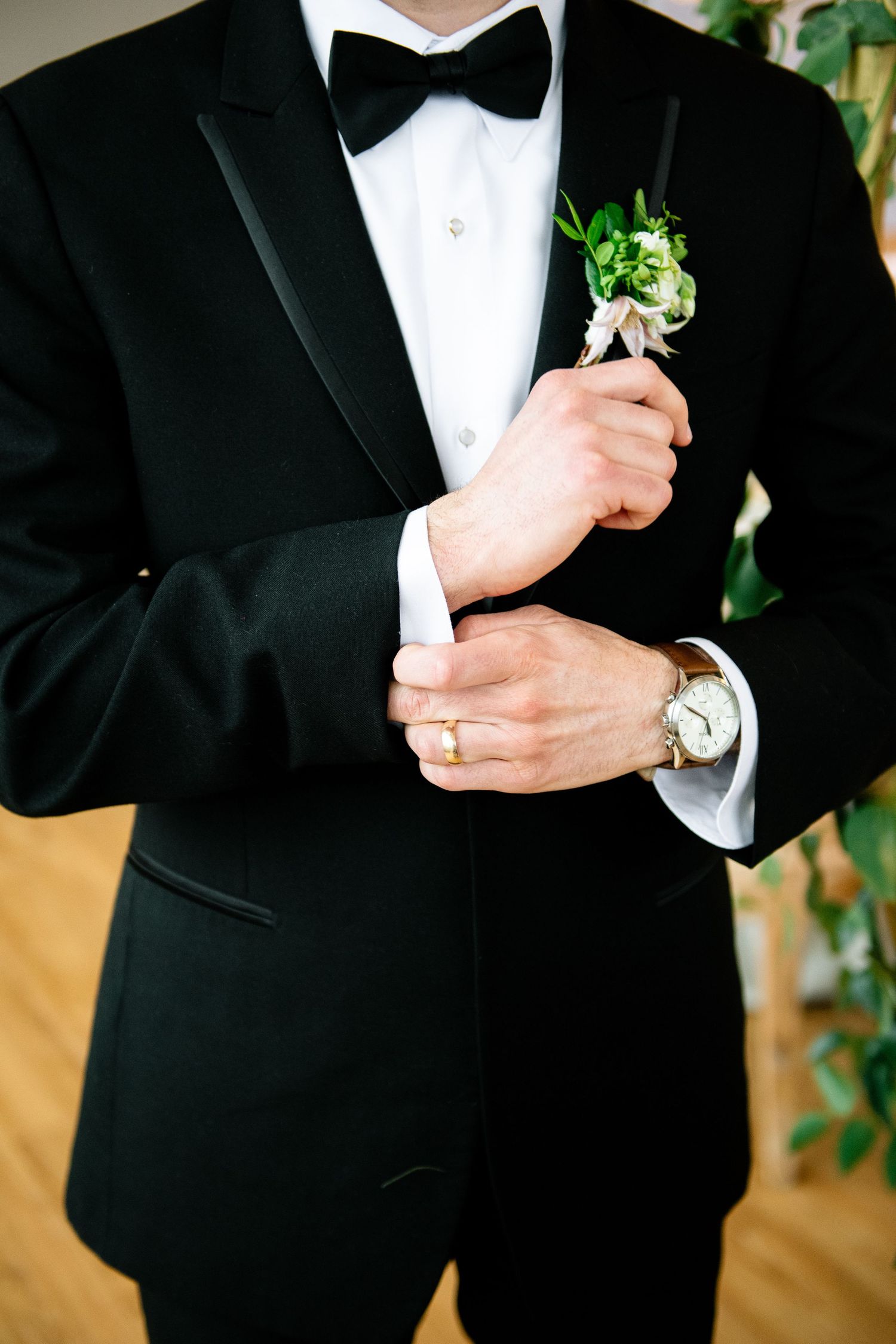 Groom in black tuxedo with bow tie adjusting cufflinks, wearing gold watch and ring, with green boutonniere on lapel.