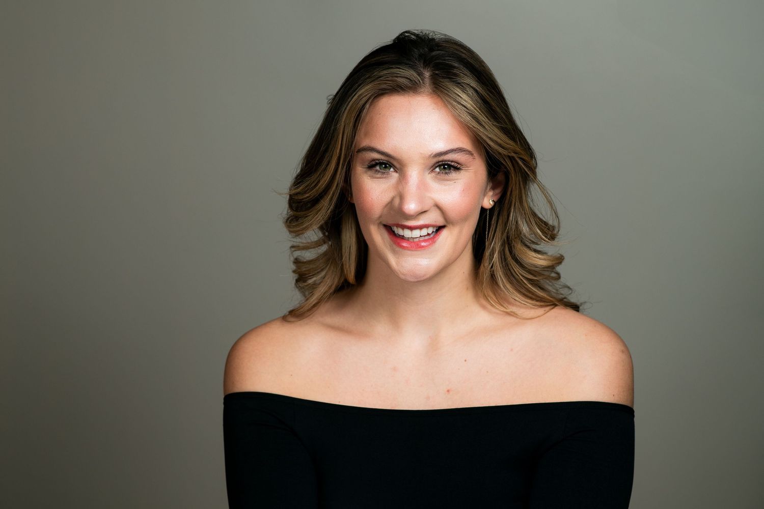 Smiling young woman with wavy brown hair wearing a black off-shoulder top against a gray studio background.