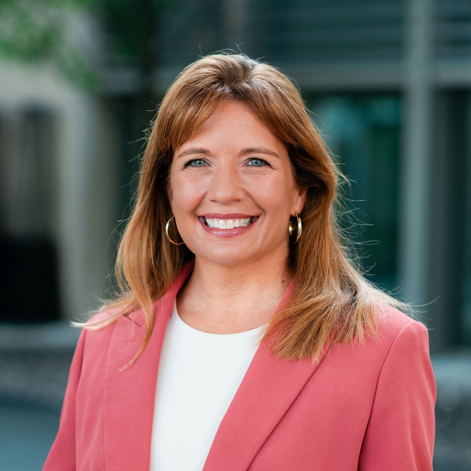 Professional headshot of salesperson in a coral blazer and white top smiling against a blurred office background.