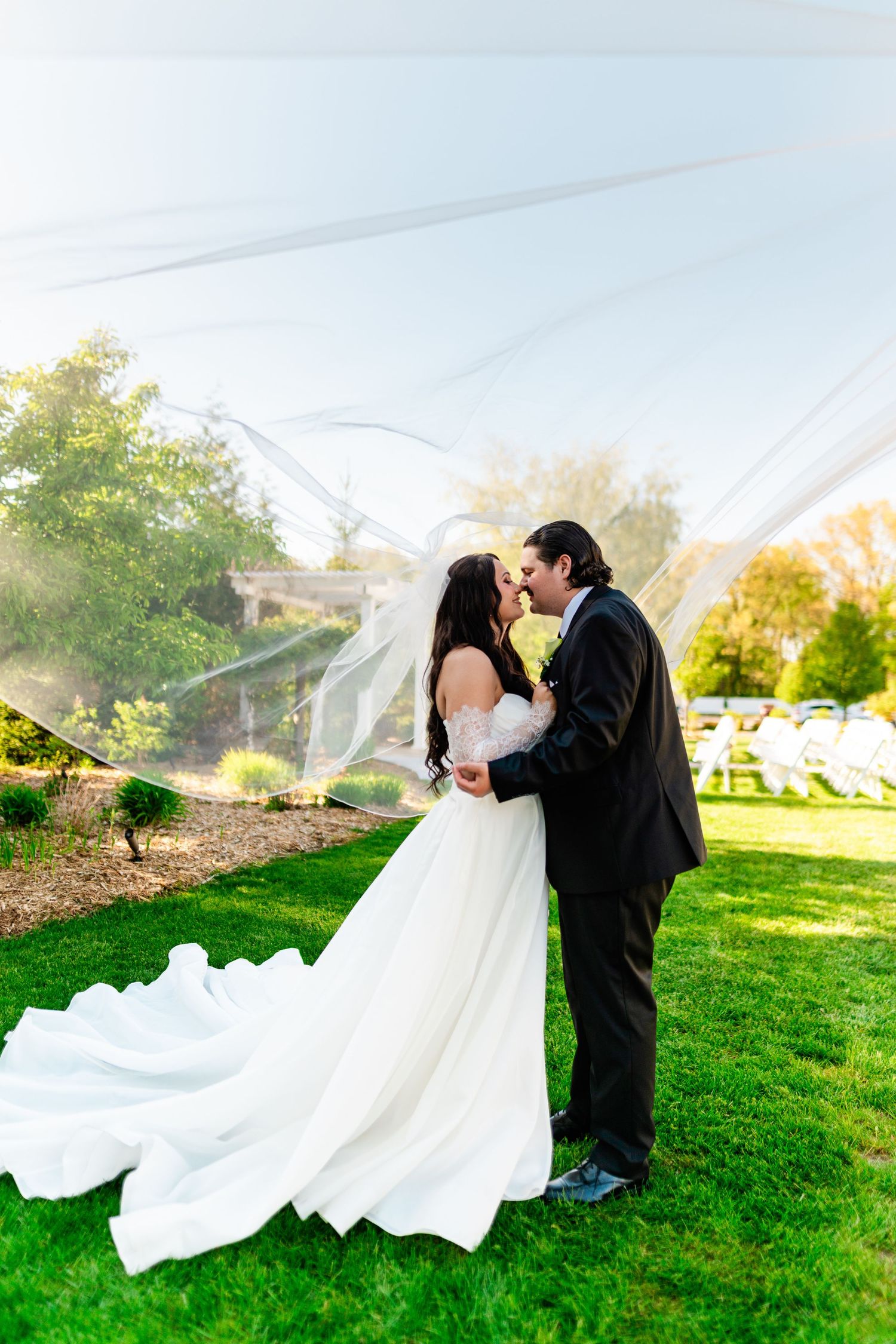 Bride and groom share a romantic kiss outdoors on green lawn, with flowing white veil billowing in the breeze.