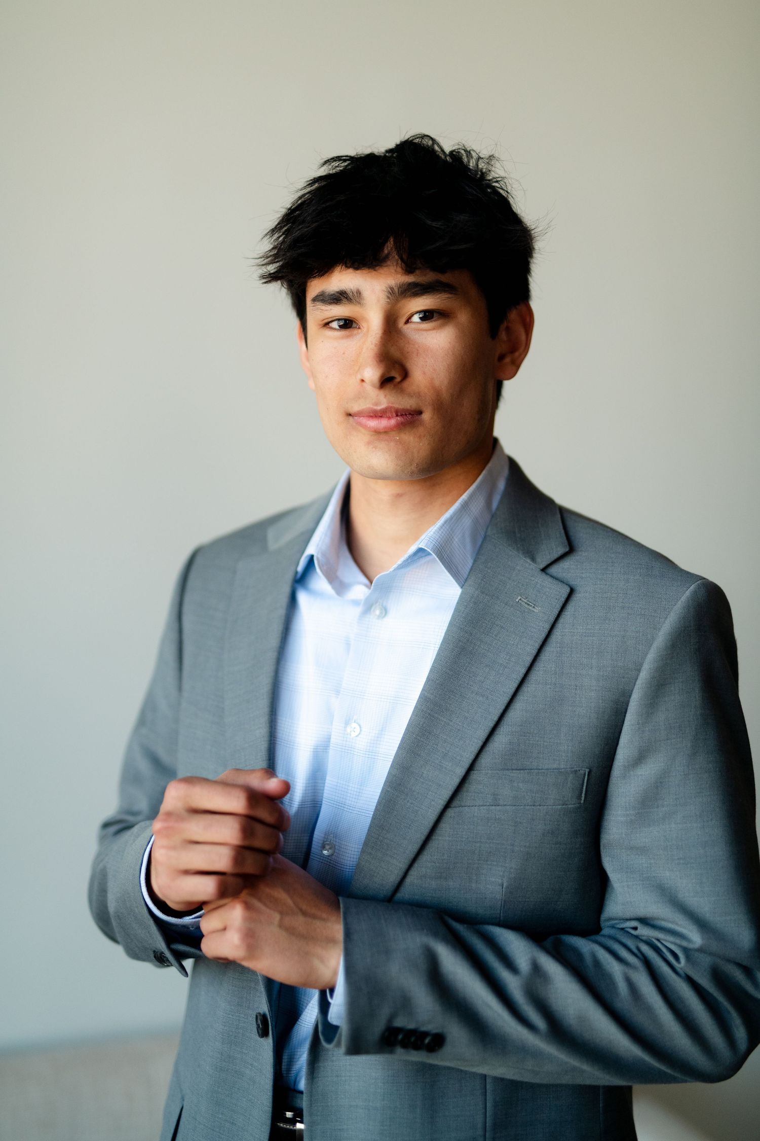 Professional model in a grey suit adjusts his collar while standing against a light background.