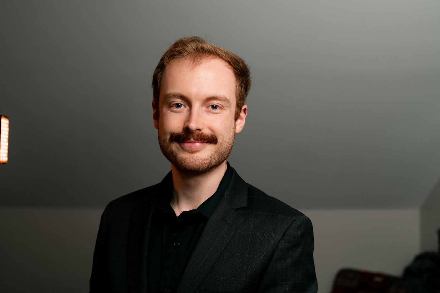 A person in a black suit jacket smiles warmly against a dark background in a professional headshot.