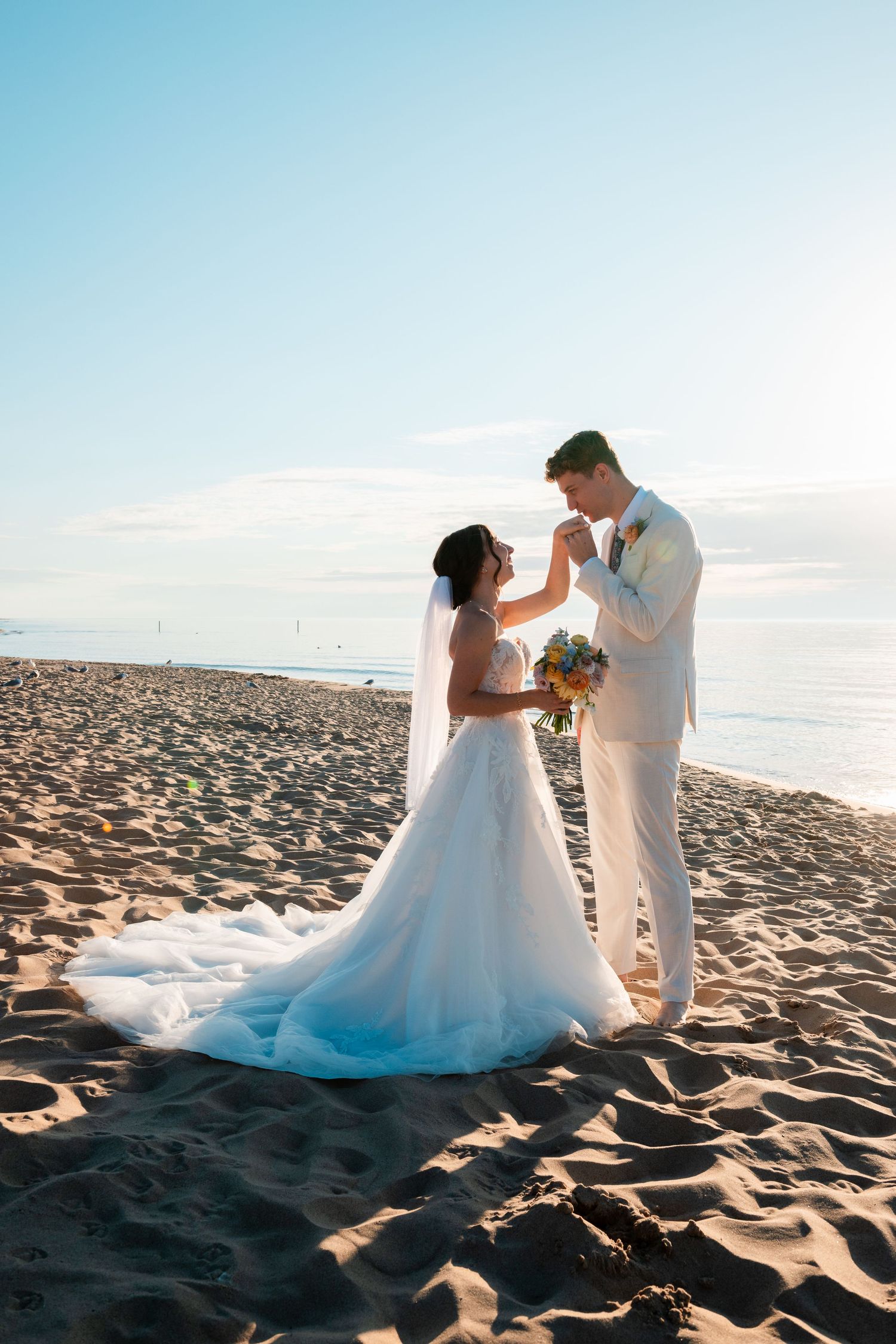 A couple dances together on a sandy beach at sunset in wedding attire.