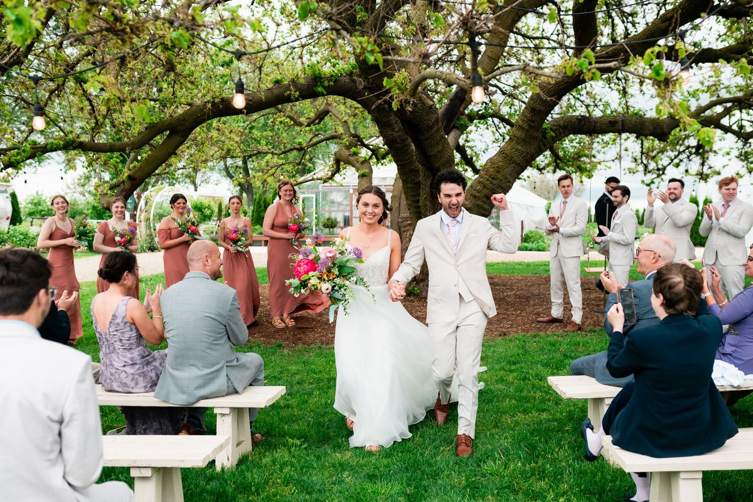 An outdoor wedding ceremony takes place under a blooming tree with white benches and pink-dressed attendees on a grassy lawn.