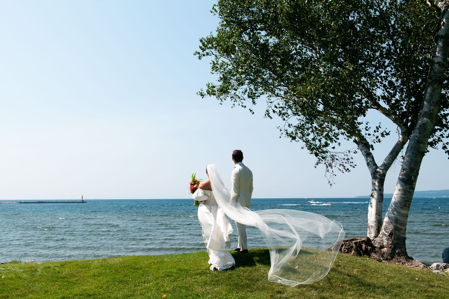 Newlywed couple in white wedding attire standing by the waterfront near a birch tree on a sunny day.