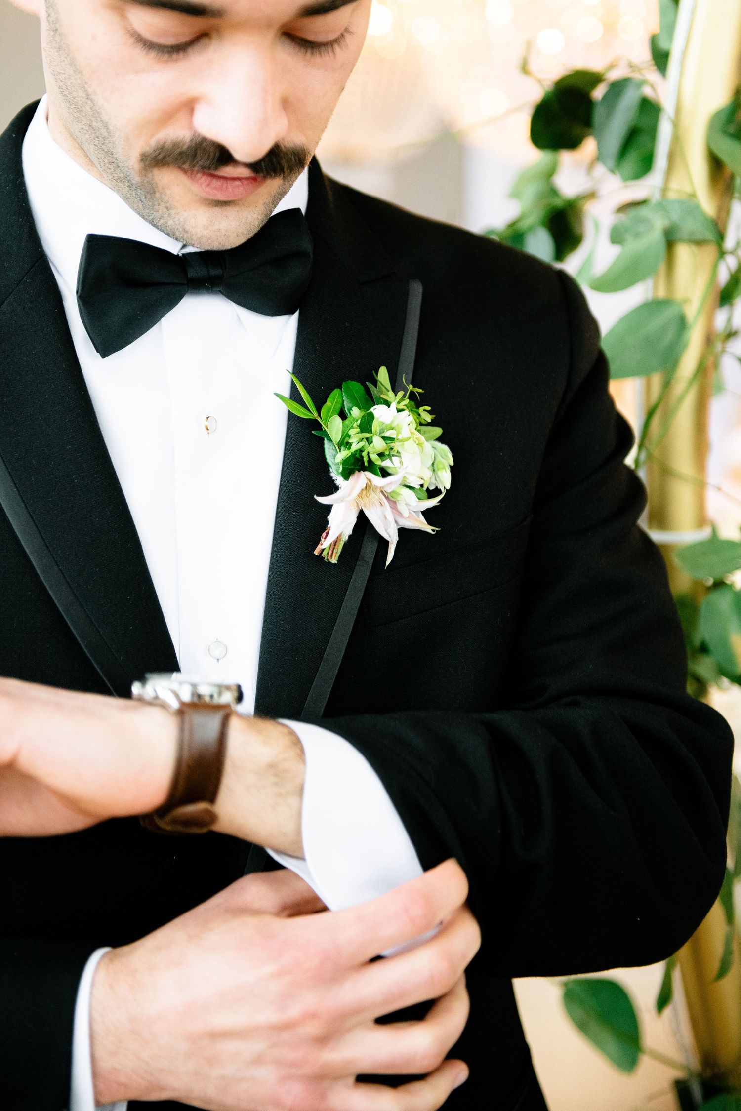 Close-up of groom in black tuxedo adjusting his white boutonniere with greenery accent.