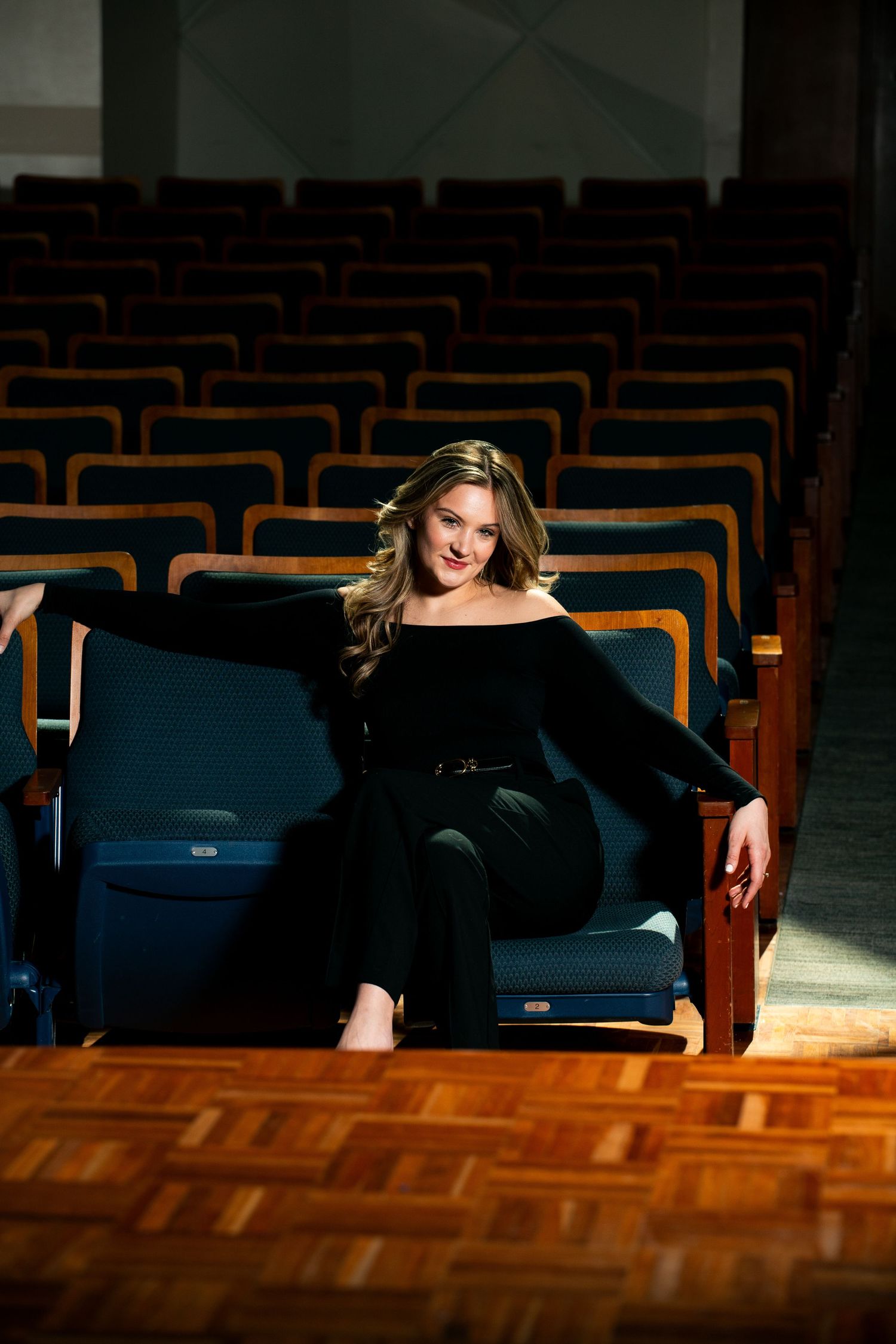 A figure in black clothing lies gracefully across wooden theater seats in dramatic lighting.