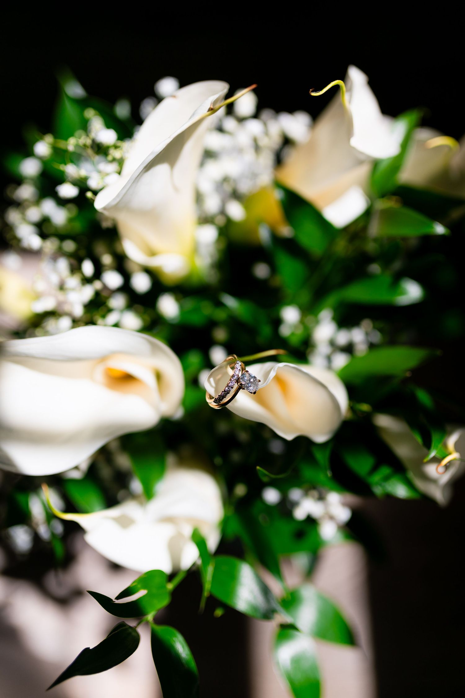 Wedding rings nestled among white calla lilies and baby's breath in a bridal bouquet against dark background.