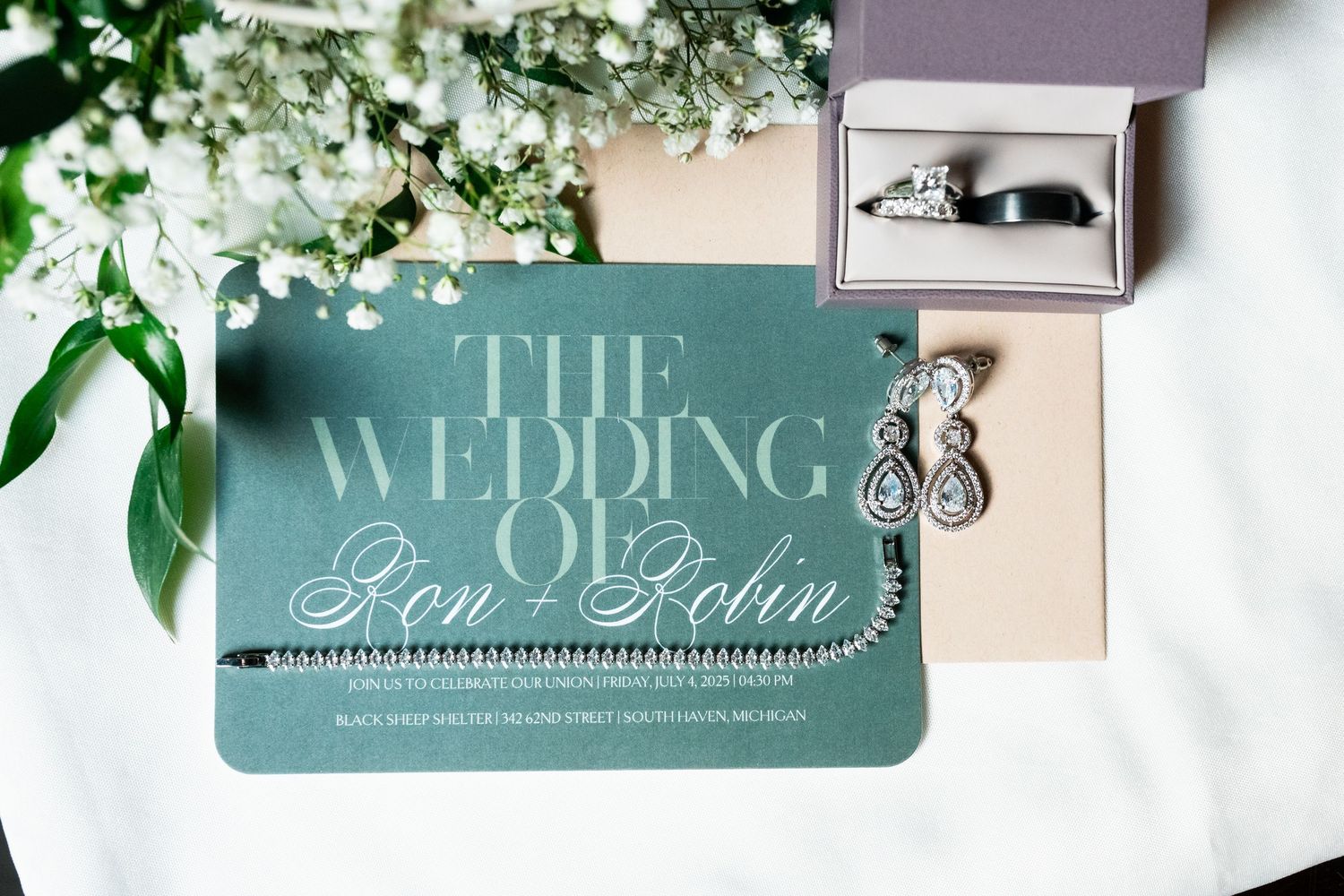 Elegant green wedding invitation for Ron and Robin displayed with diamond earrings and wedding rings in a box.
