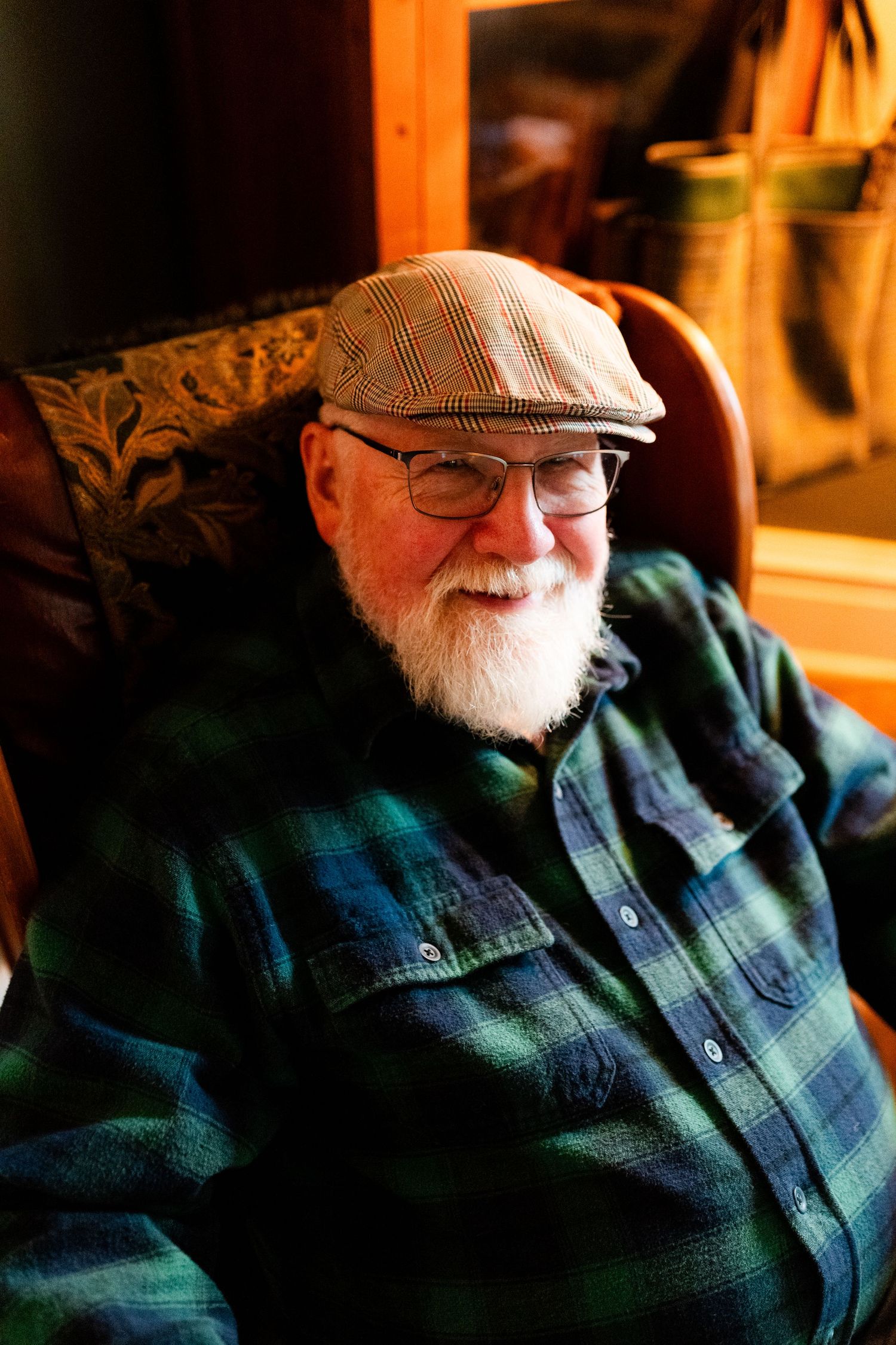 A person wearing a plaid flannel shirt and flat cap sits in a chair with a warm smile.