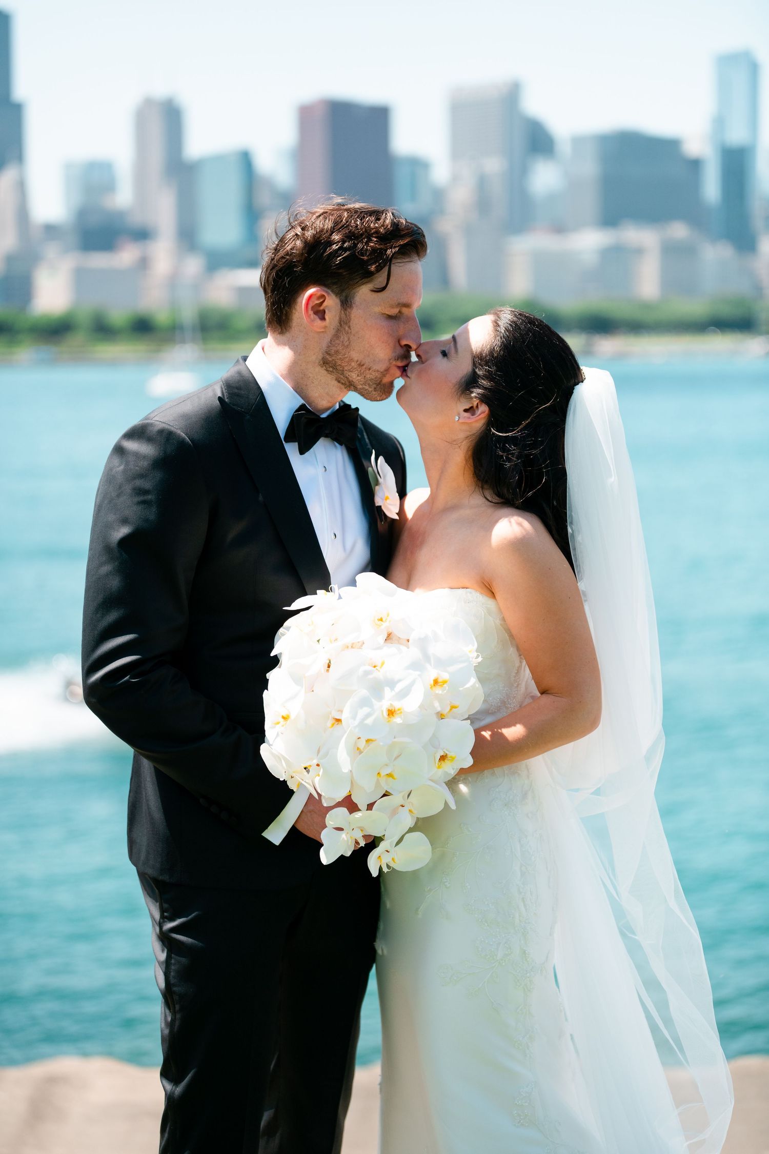 Bride and groom share a kiss by the waterfront with Chicago skyline backdrop, bride holding white orchid bouquet.