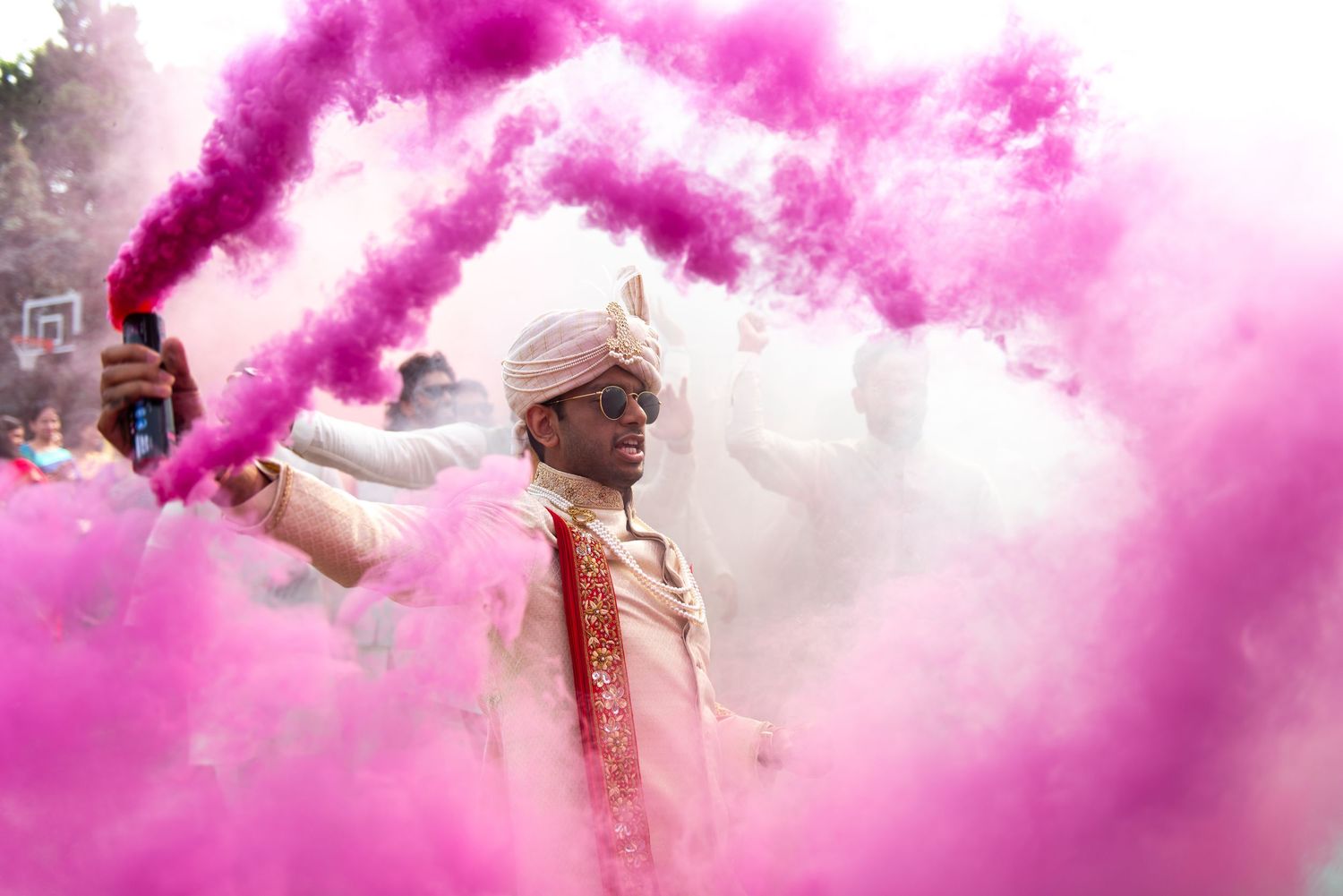 Pink smoke fills the air during a vibrant outdoor celebration with people gathering beneath cloudy skies.