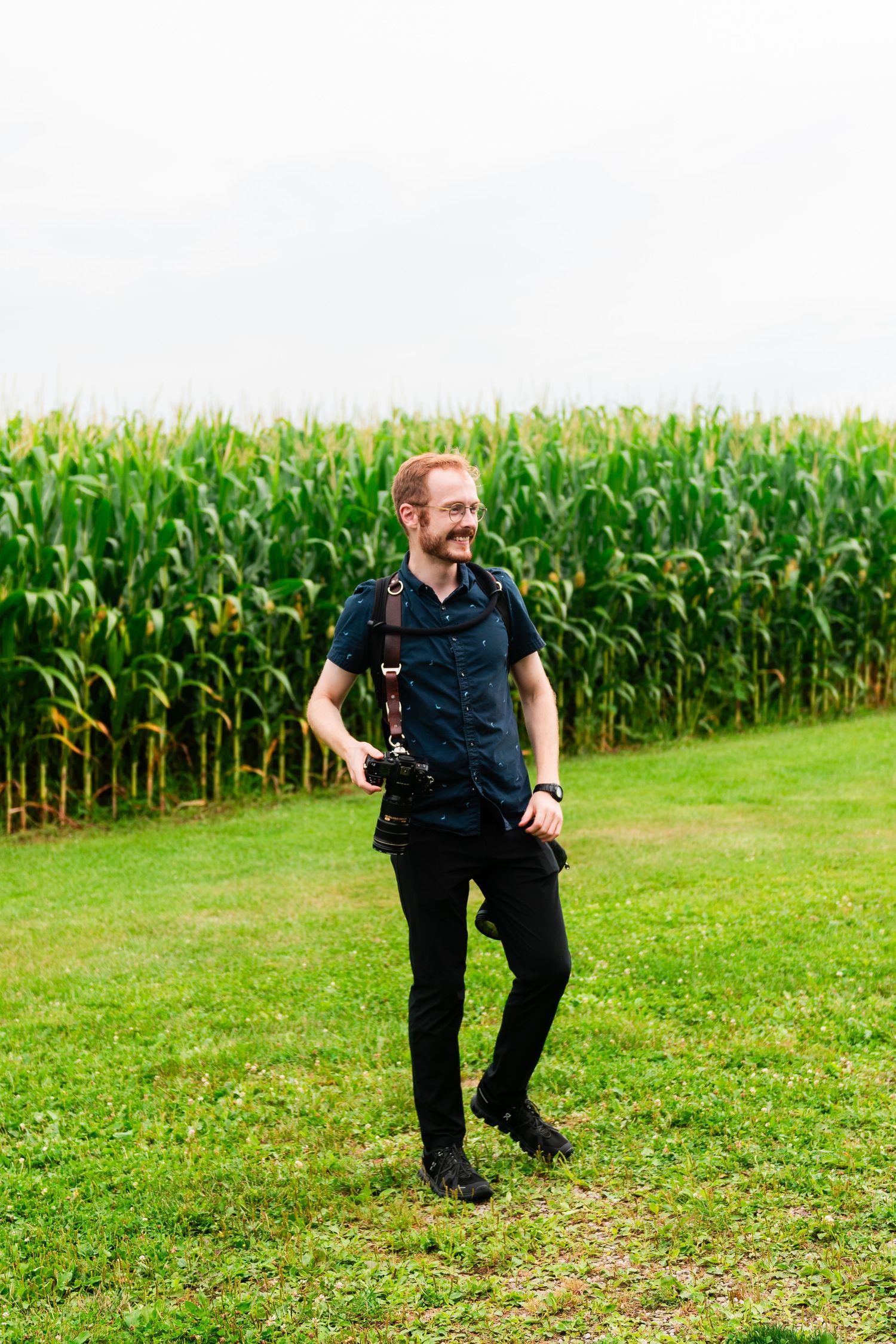 Person in dark clothing runs playfully across a grassy field next to tall corn stalks on a sunny summer day.