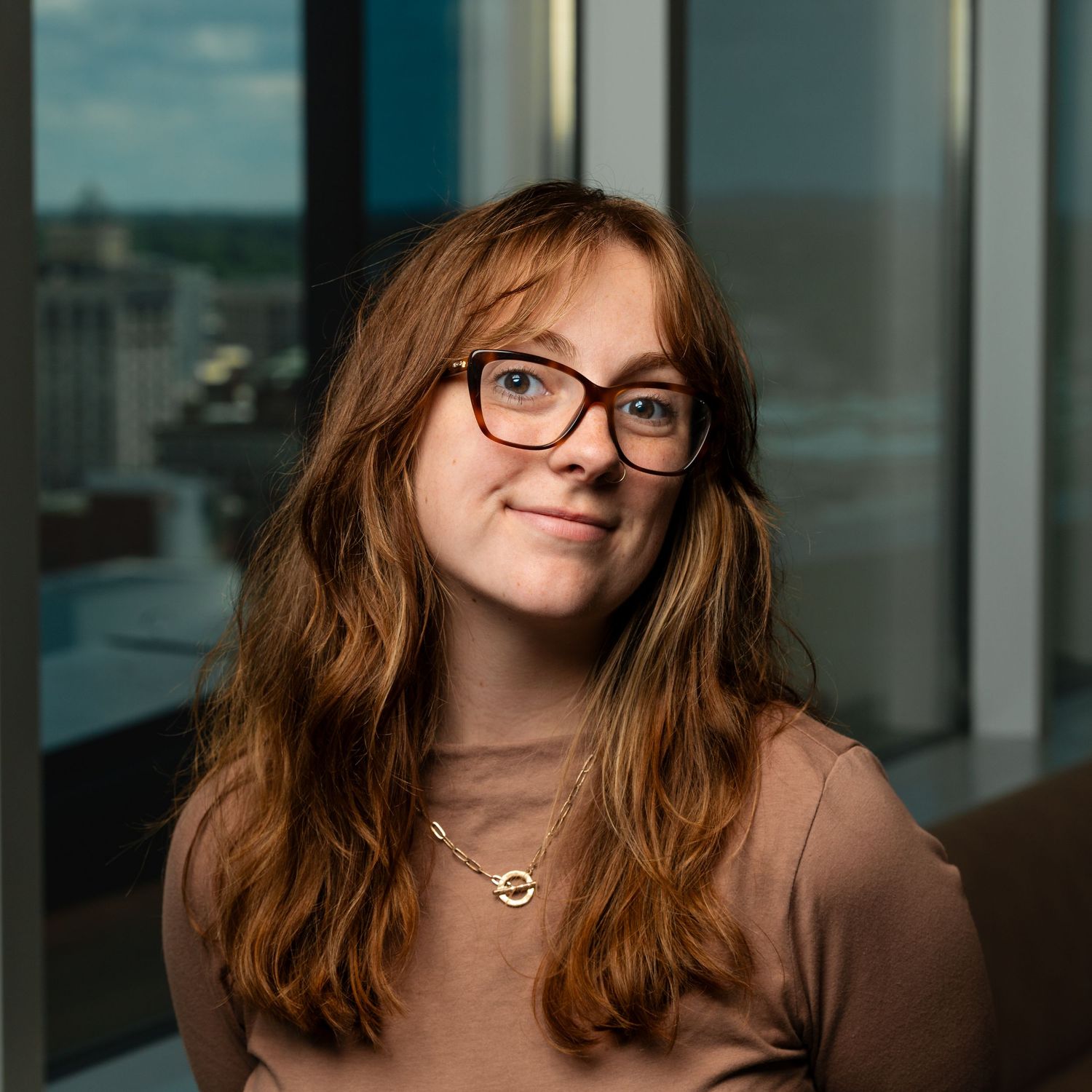 Young woman with red hair and glasses smiling for a headshot indoors near a window with a grand rapids background.