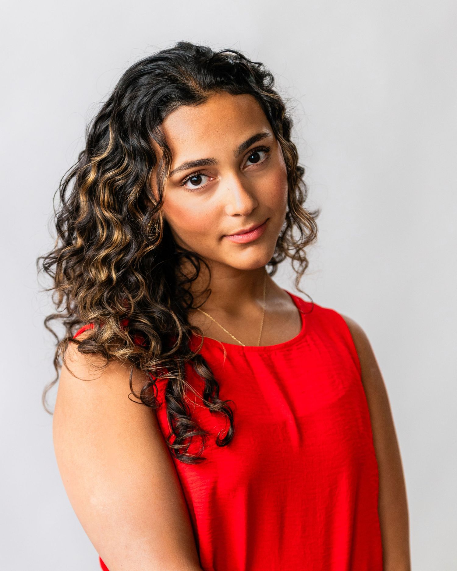 Young woman with curly highlighted hair wearing a red sleeveless top poses against a light gray background.