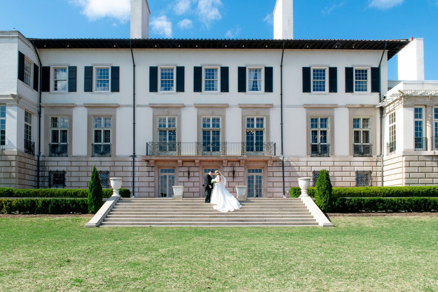 Elegant white mansion with grand stone staircase and manicured lawn features classic colonial architecture and black shutters.