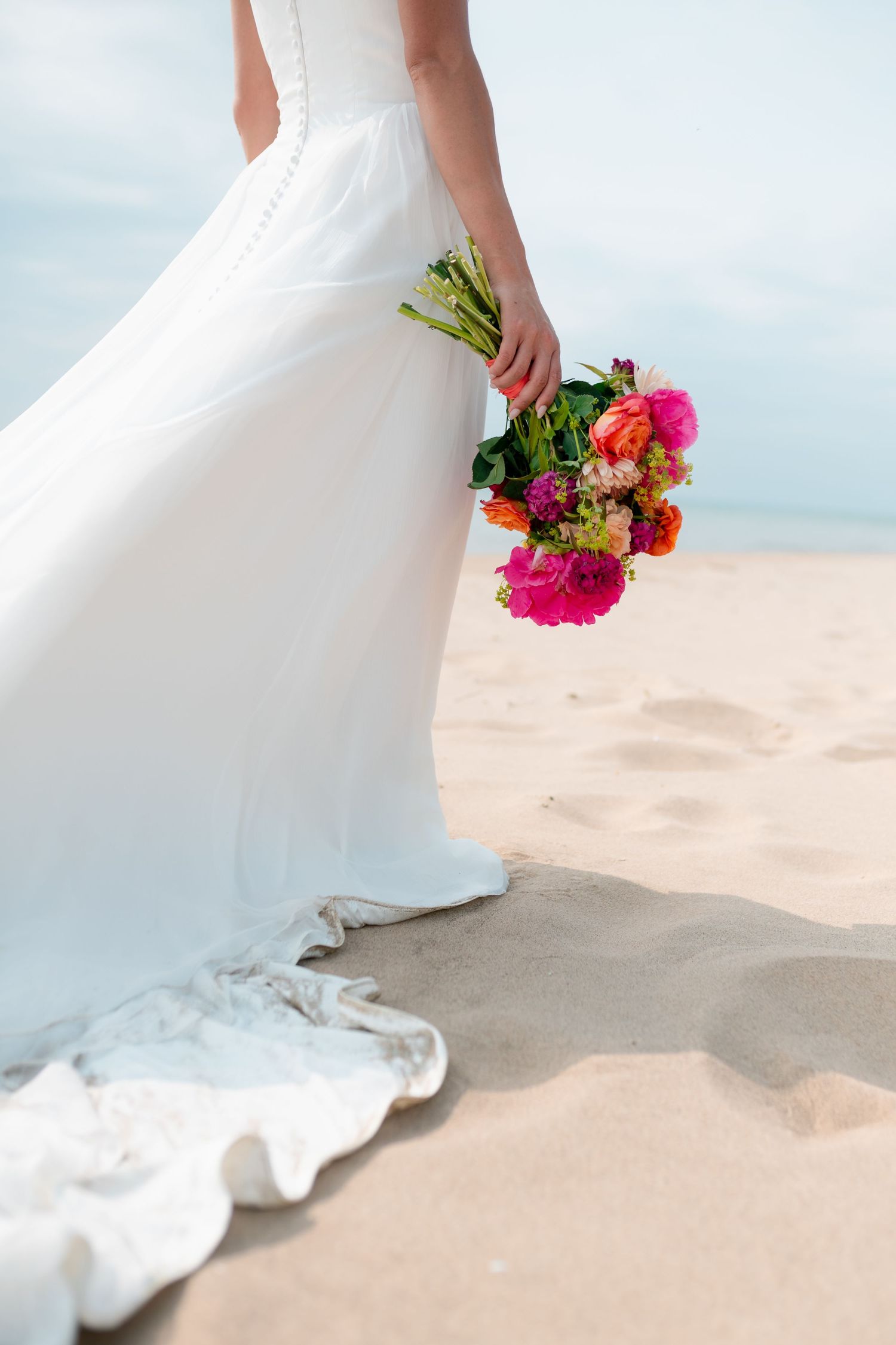 Close-up view of a white wedding dress train dragging in beach sand with a vibrant floral bouquet peeking into frame.