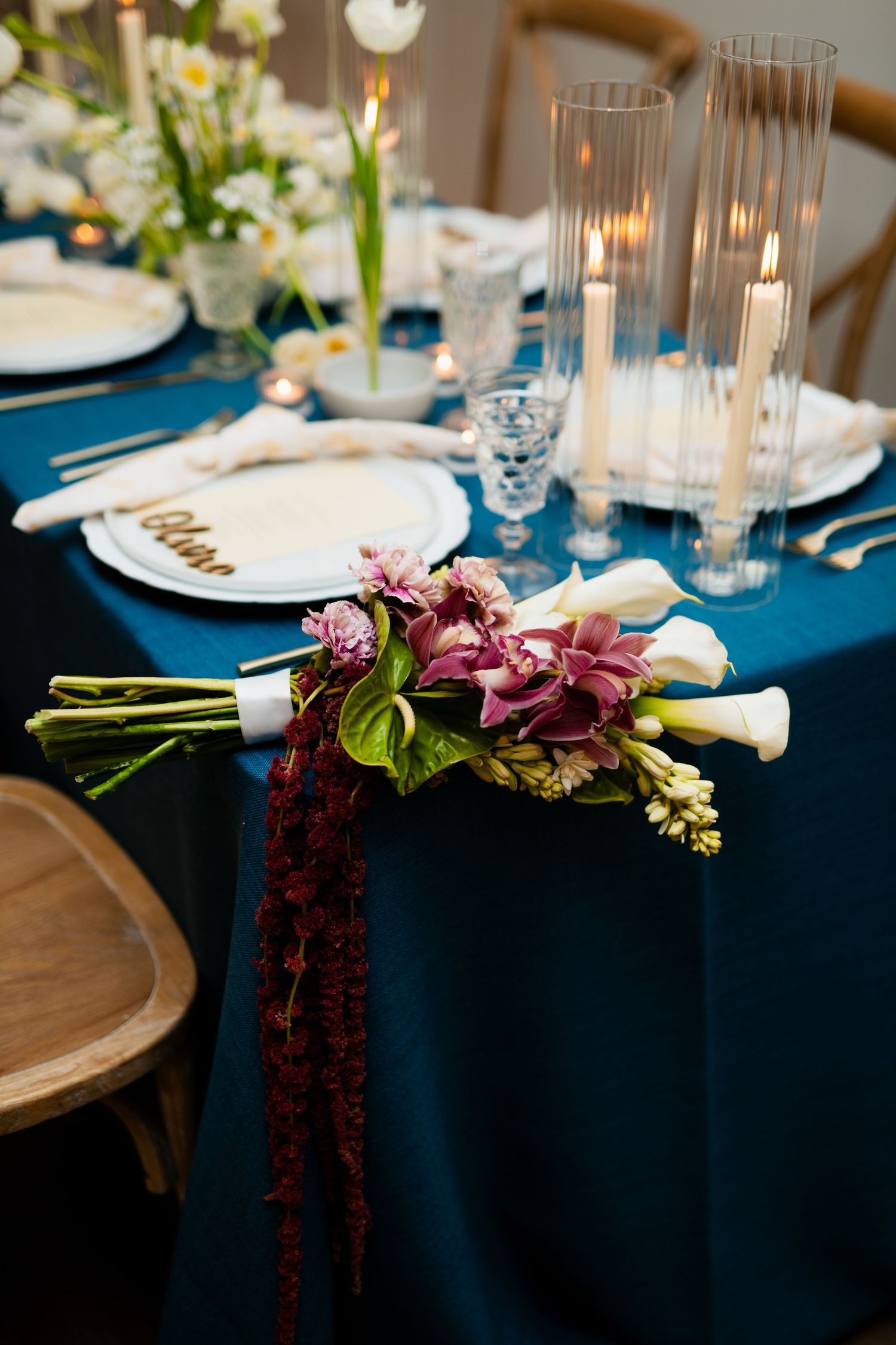Elegant wedding table with navy blue linen, purple floral bouquet, tall candles, and white plates with gold accents.