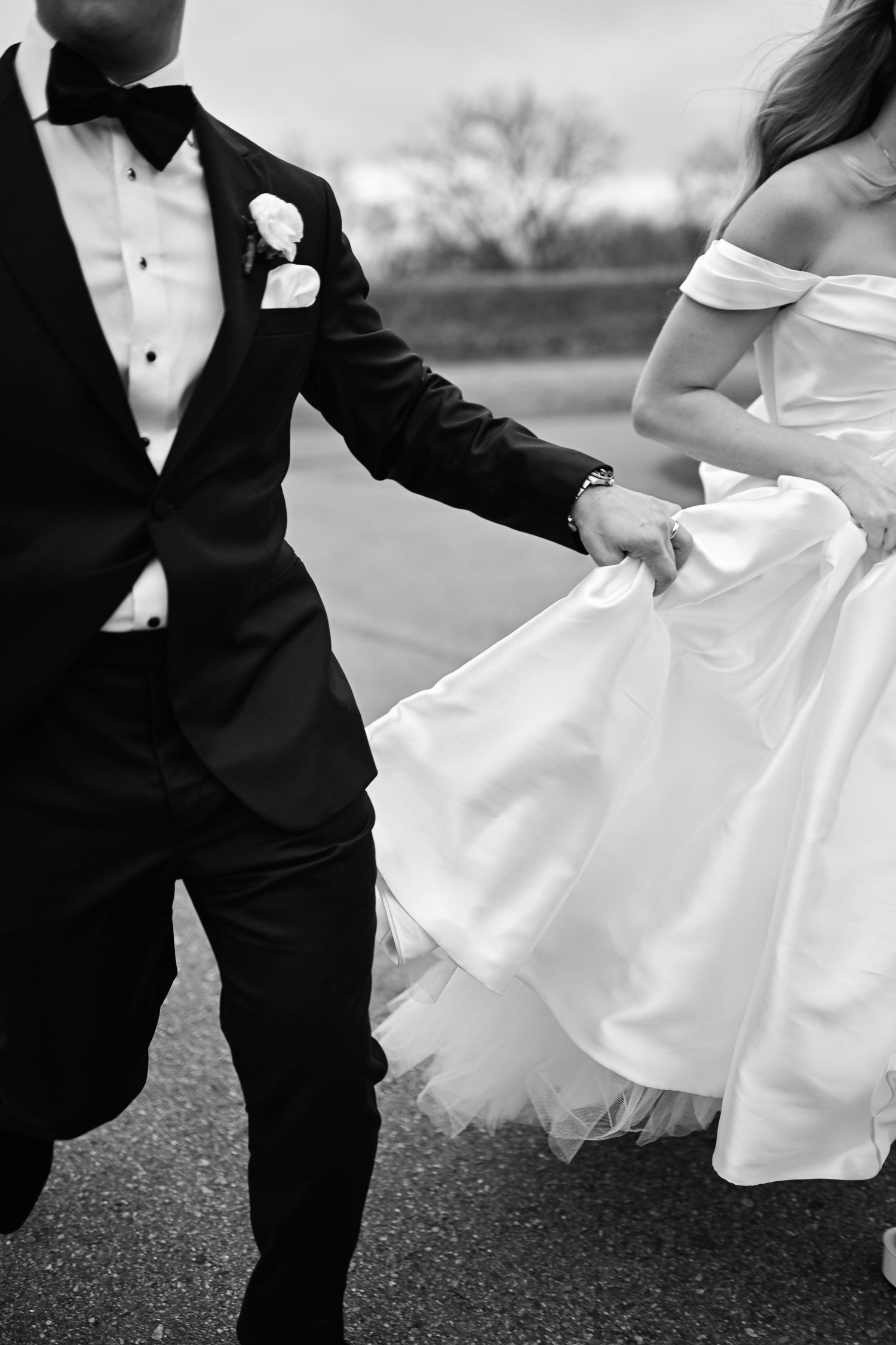 Black and white photo of bride and groom holding hands, walking in wedding attire, tuxedo and off-shoulder gown.