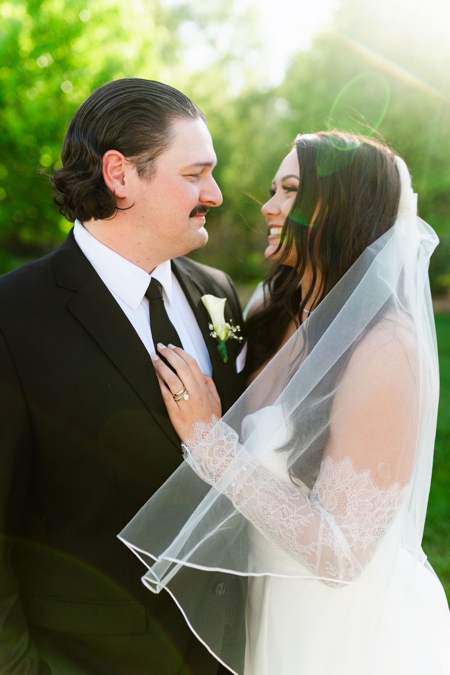 Bride and groom share a romantic moment outdoors, bride in lace veil and groom in black suit with boutonniere.