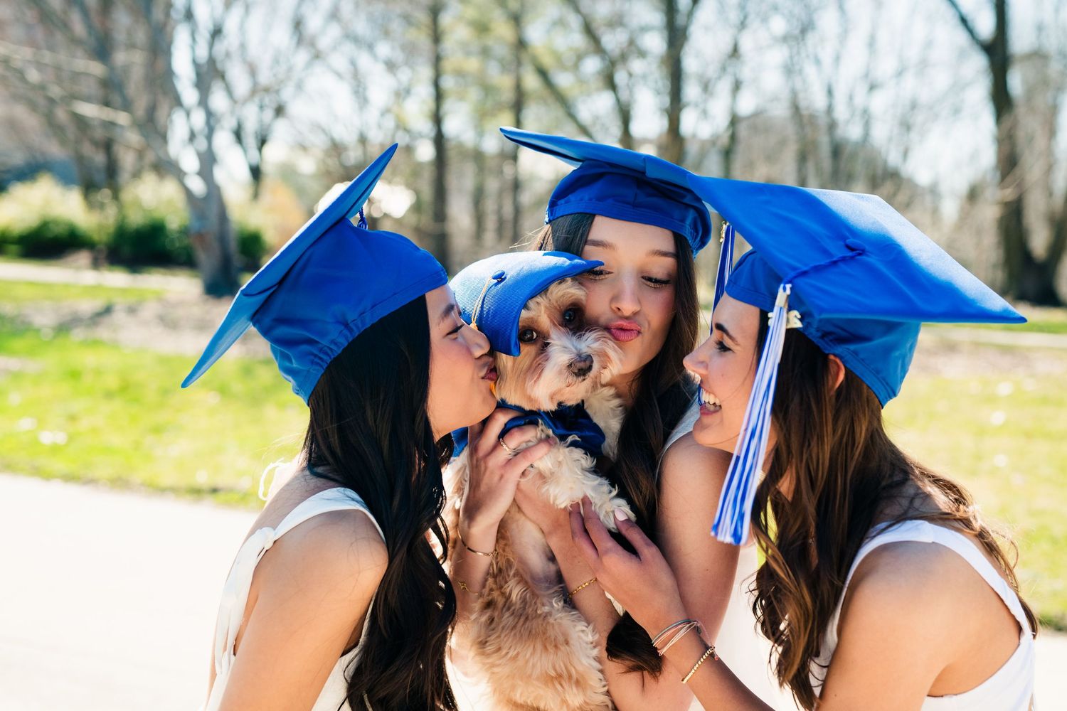 Two graduates in blue caps and white outfits share happy kisses with a small brown dog outdoors on a sunny day.
